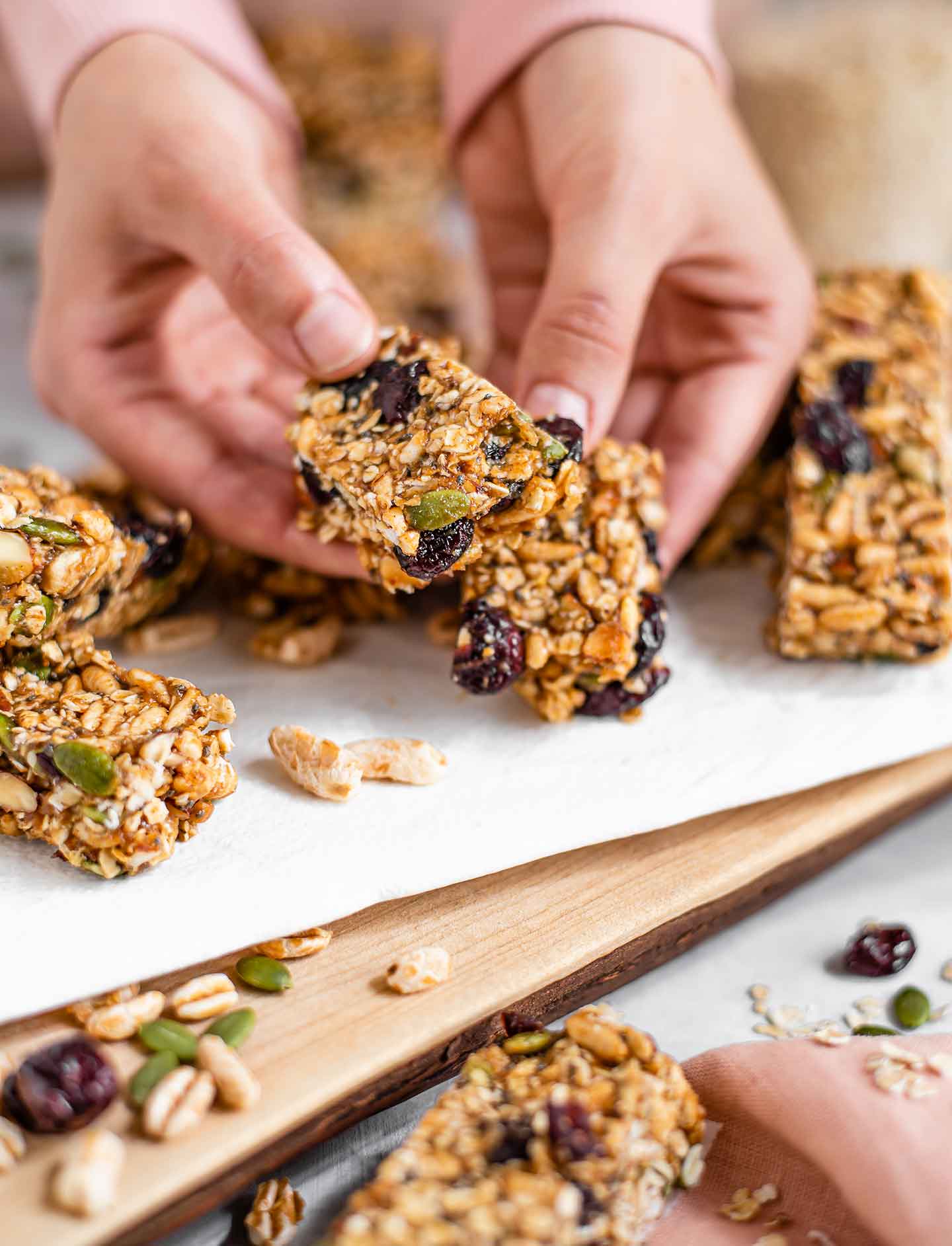 Side view of two hands breaking apart a puffed rice granola bar to show the insides. The granola bars are thick and filled with puffed grains, oats, almonds, and festive pumpkin seeds, and dried cranberries.