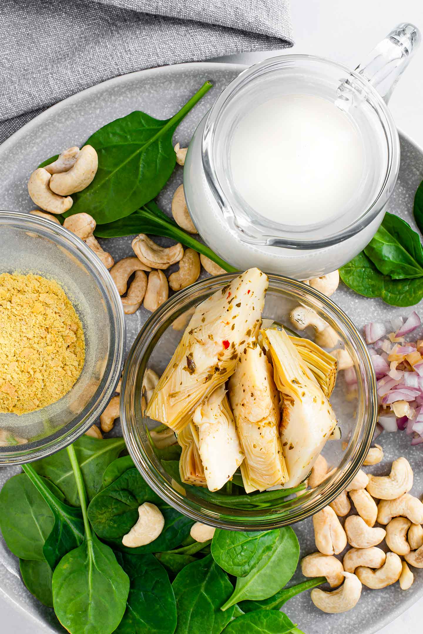 Top down view of artichoke hearts, cashews, milk, nutritional yeast, onion, and spinach on a tray.