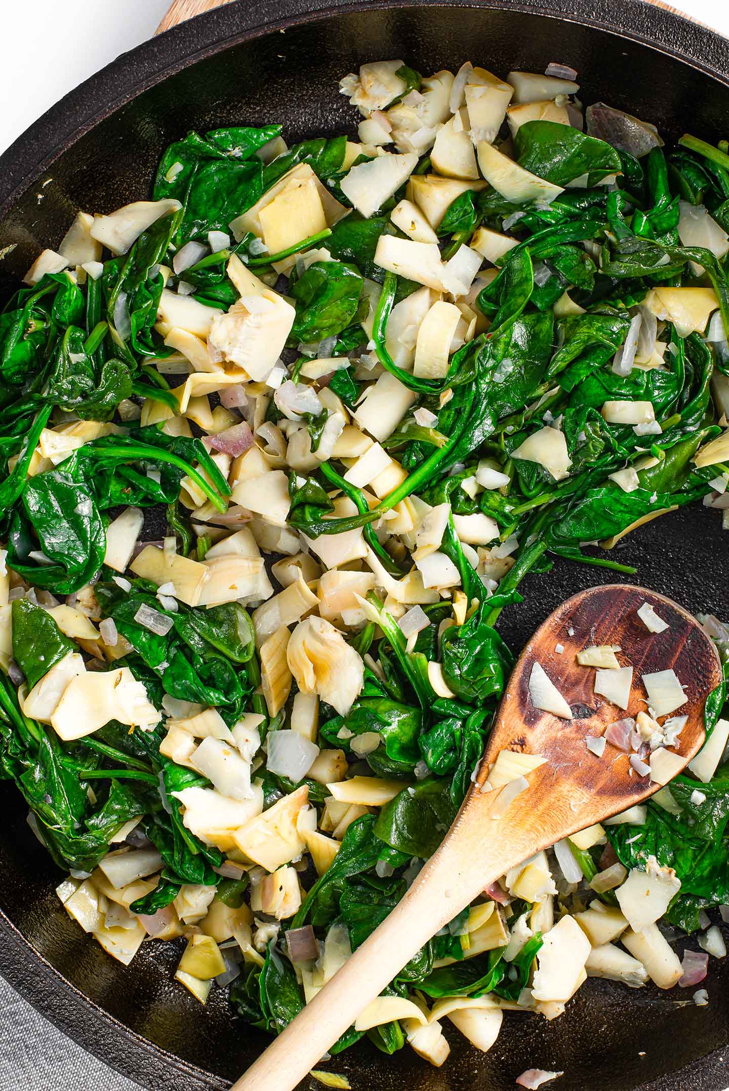 Top down view of wilted spinach and chopped artichoke hearts in a skillet with a wooden spoon.