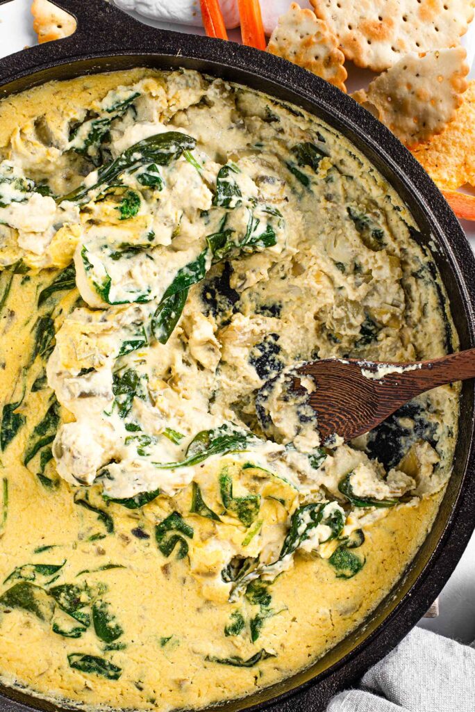 Top down view of creamy spinach artichoke dip in a cast iron skillet with a bamboo spoon digging into the mixture.