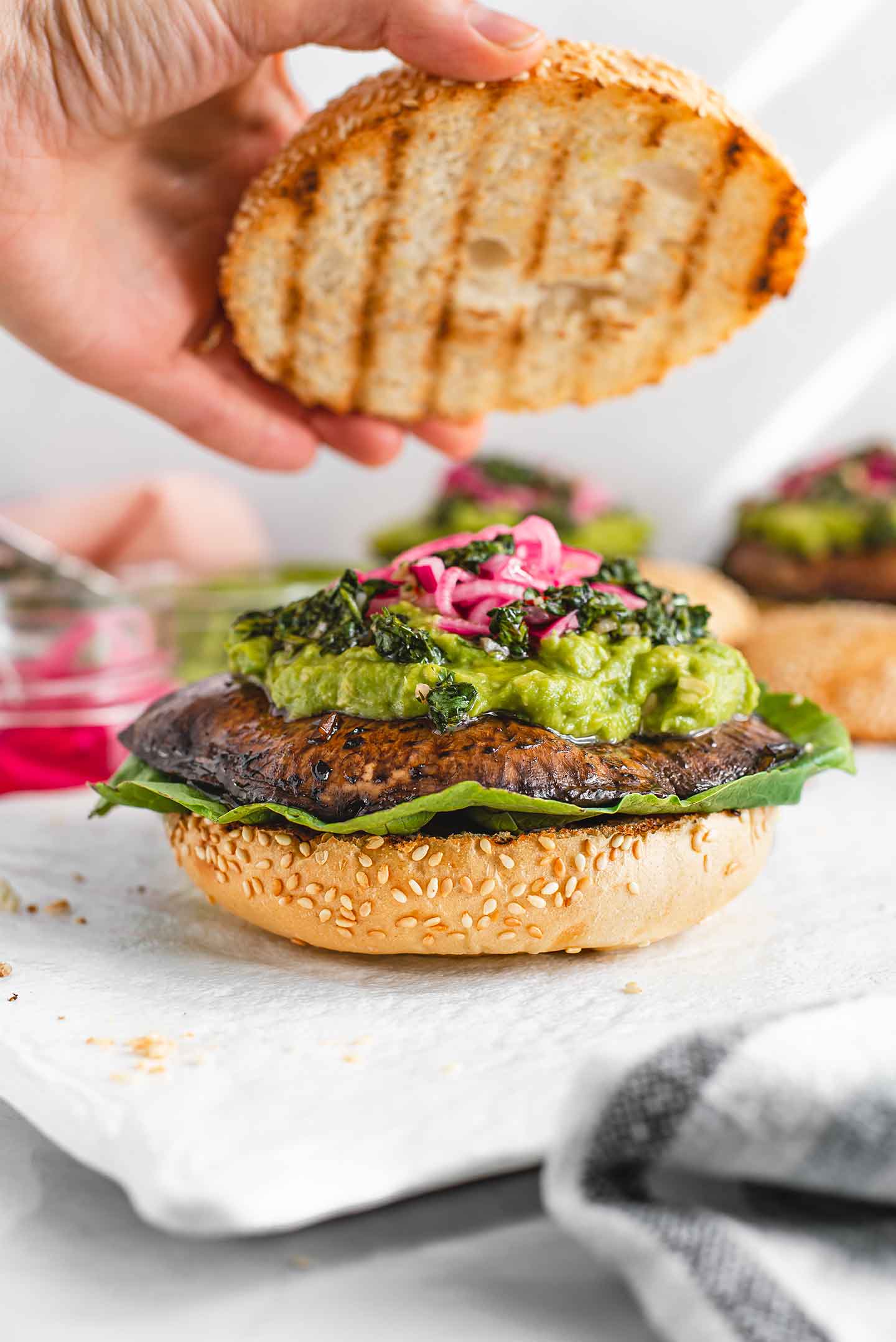 Side view of a hand placing the top bun on an avocado chimichurri portobello burger.