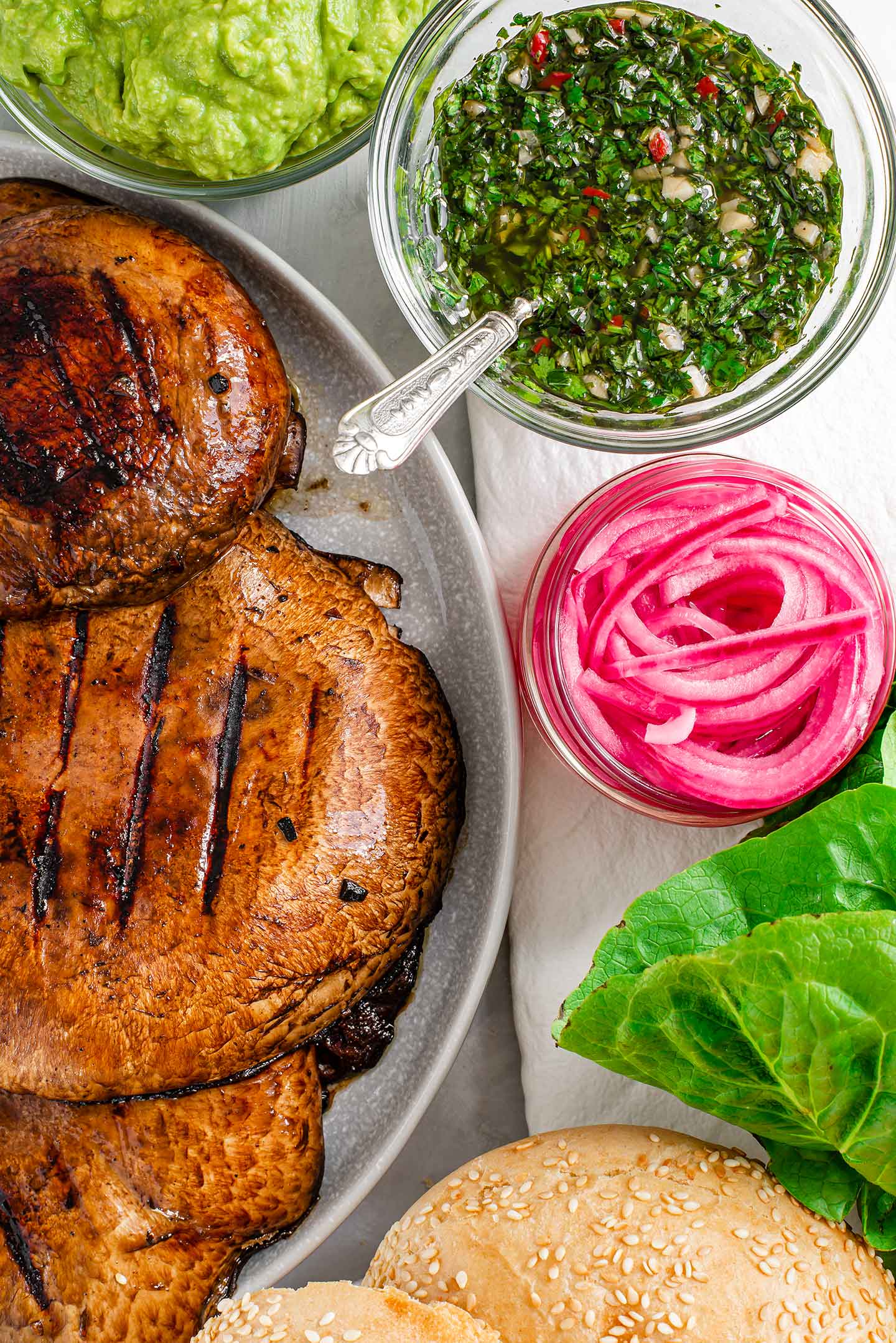 Top down view of portobello mushrooms with grill marks, mashed avocado, chimichurri, pickled red onion, romaine lettuce, and sesame burger buns.