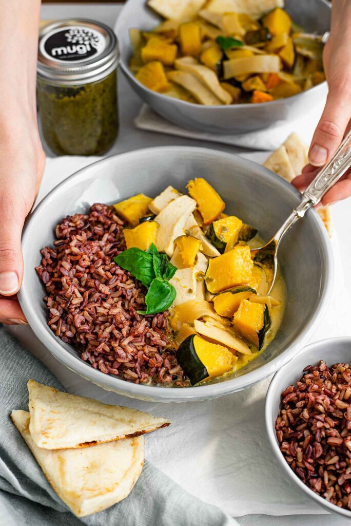 Hands place a bowl of Thai green curry in the frame. The squash curry is served with wild rice and Mugi green curry paste is in the background.