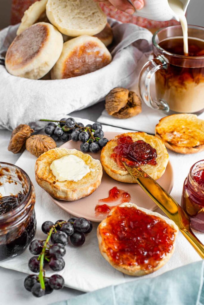 Side view of toasted Portuguese sweet muffins buttered and smeared with jam. Milk is poured into coffee in the background, a bread basket overflows with muffins and grapes and figs surround the cut muffins.