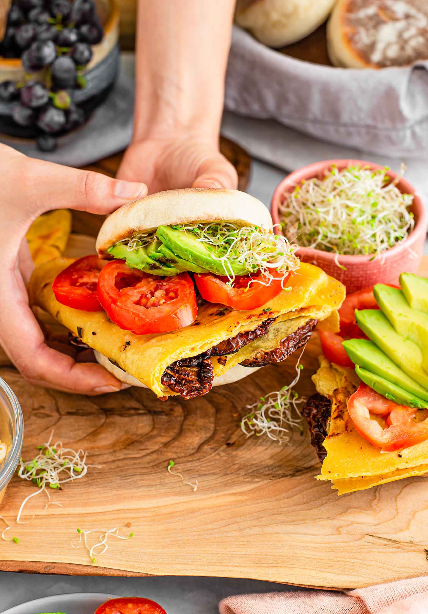 Side view of two hands picking up a loaded vegan Portuguese breakfast sandwich. Ingredients and another partially constructed sandwich scatter the wooden board. 