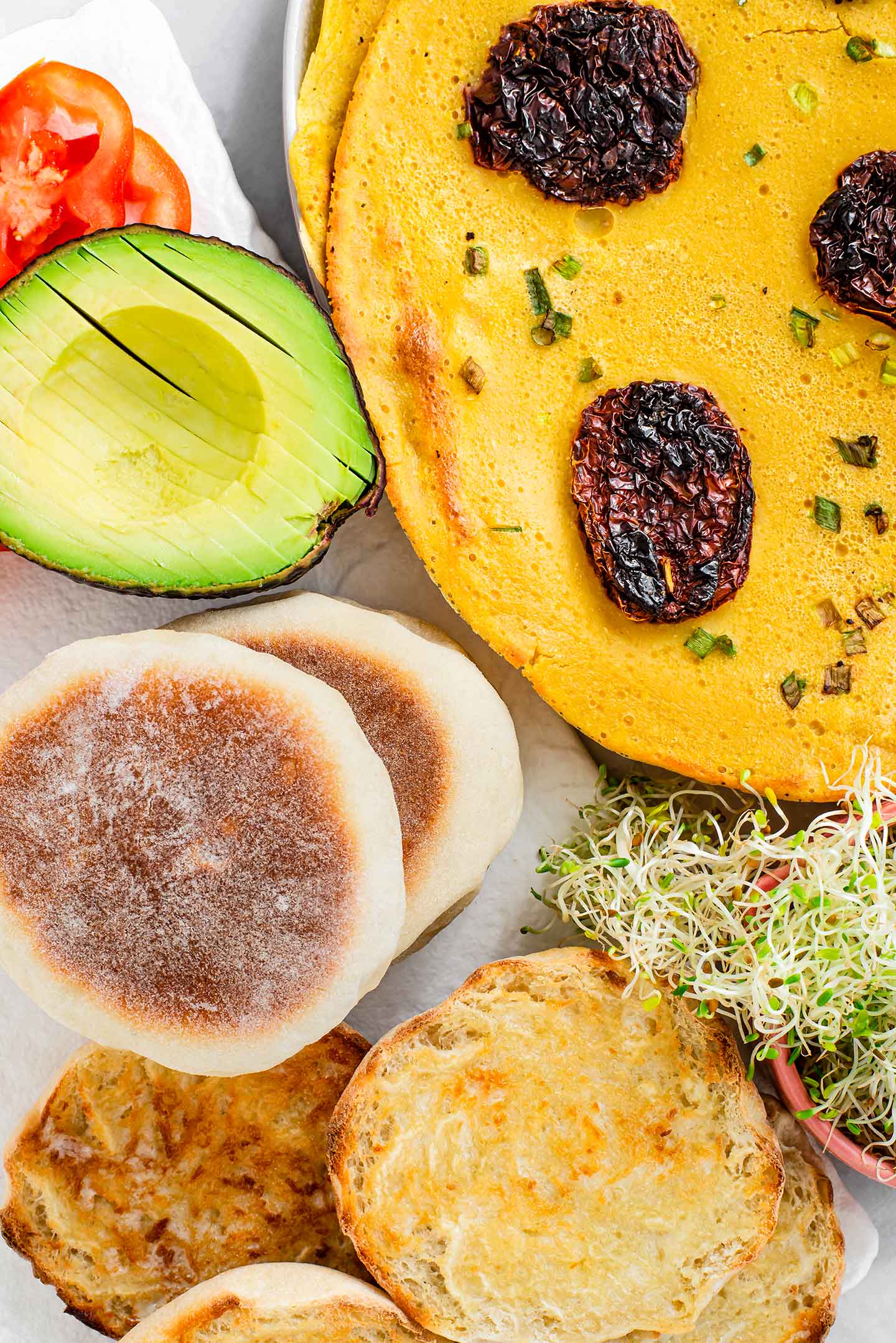 Top down view of a chickpea omelette with sun-dried tomato chourico beside bolos levedos, a sliced avocado, tomato, and sprouts.