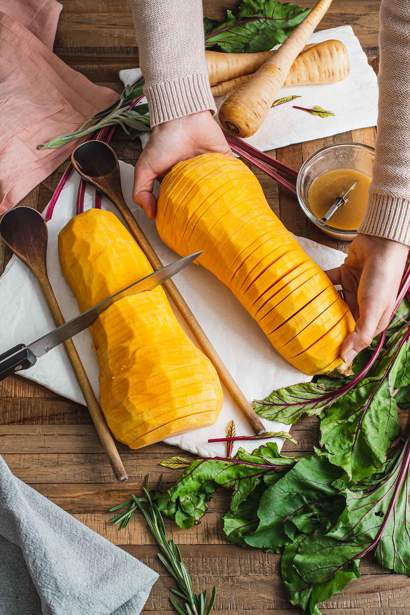Top down view of a butternut squash being cut hasselback style. Two wooden spoons lay on either side of one half of the squash and a knife rests in a slit in the centre. Hands hold up the other half of the squash, showing the thin cuts and hasselback look.