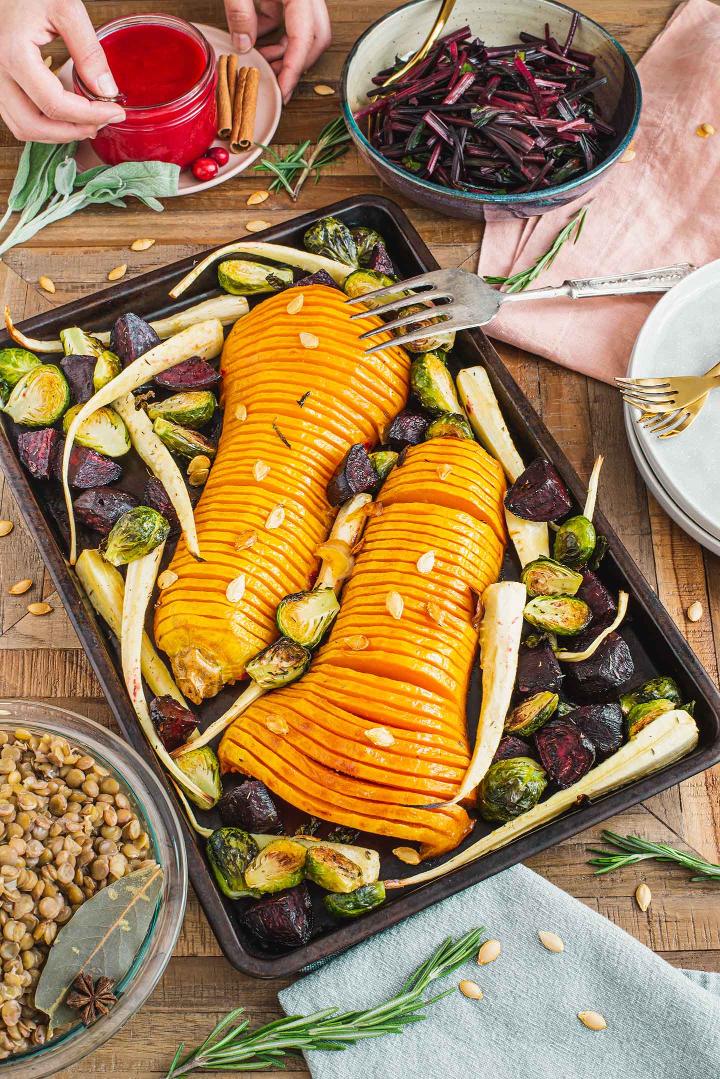 Top down view of a roasted hasselback butternut squash on a festive tray with parsnips, beets, and brussels sprouts. Lentils, sauteed beet greens, and cranberry sauce surround the tray.