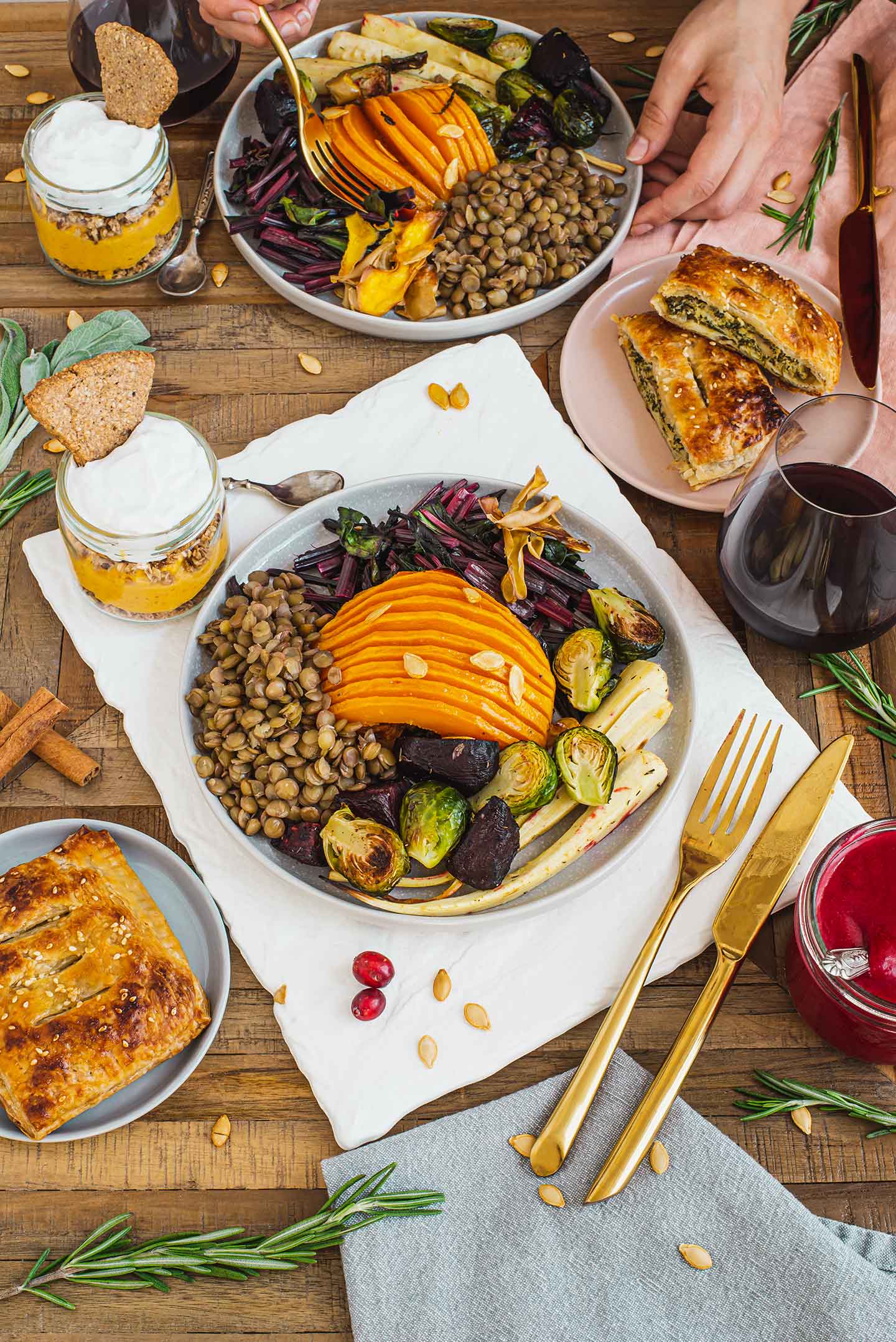 Top down view of a holiday table. Hasselback butternut squash is in the middle of a plate of roasted vegetables, beet greens, and lentils. Cranberry sauce, a puff pastry appetizer, and a pumpkin pie parfait, and a glass of red wine surround the plate.