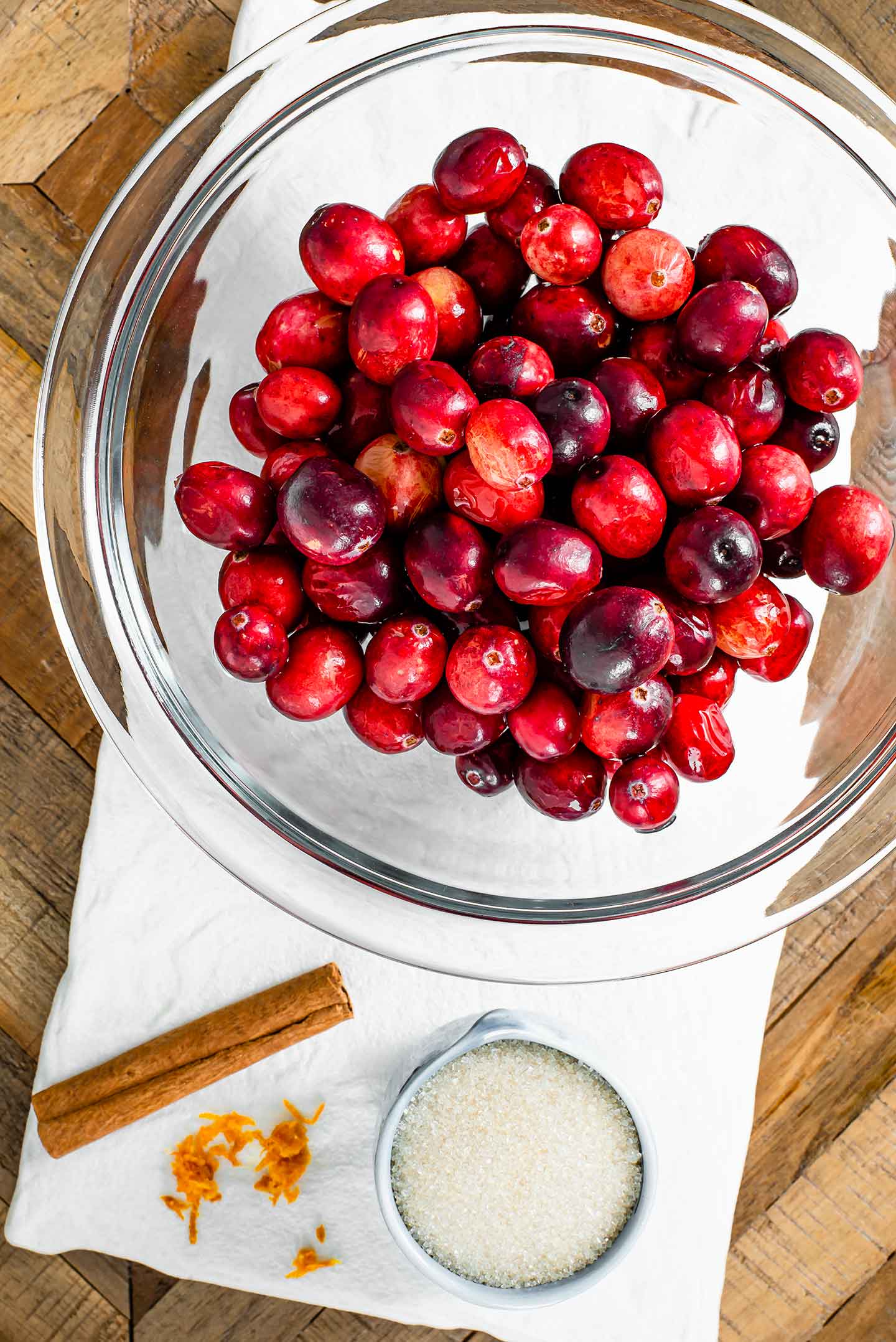 Top down view of fresh cranberries in a glass bowl. A small dish of cane sugar, some orange zest, and a cinnamon stick lay on a white tray atop a wooden table.