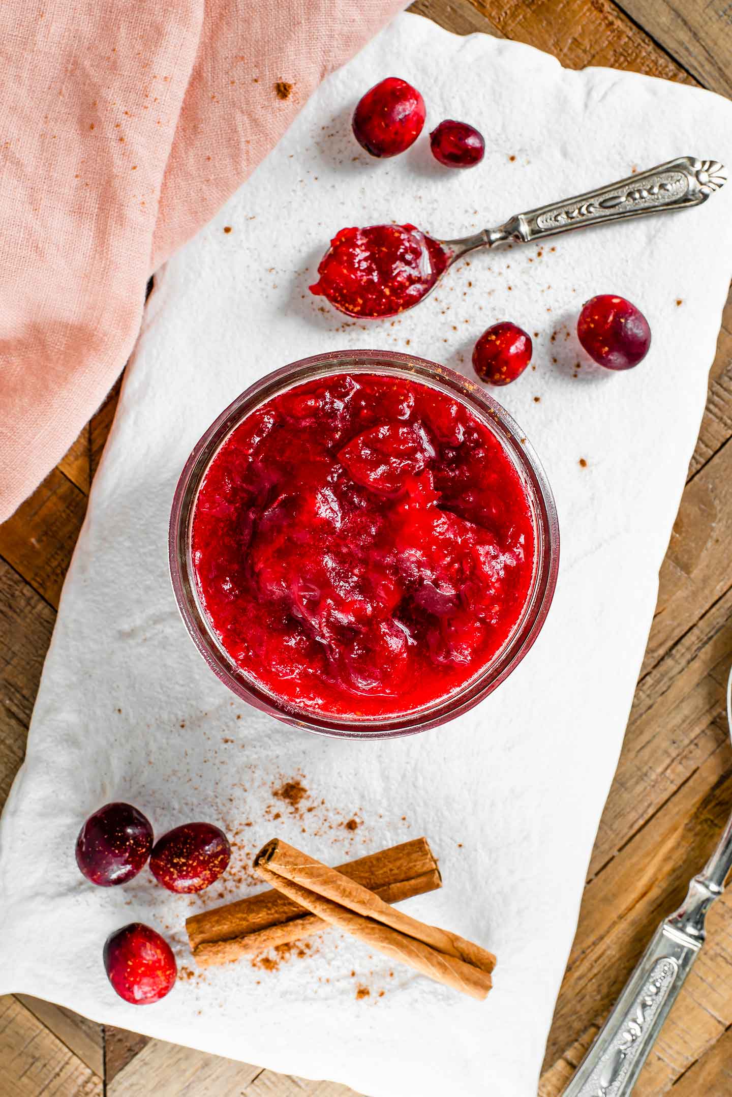 Top down view of deeply red cranberry sauce filling a glass jar. The sauce is thick with the cooked cranberries nicely softened and mashed. Cinnamon sticks and fresh cranberries surround the glass jar on a white tray.