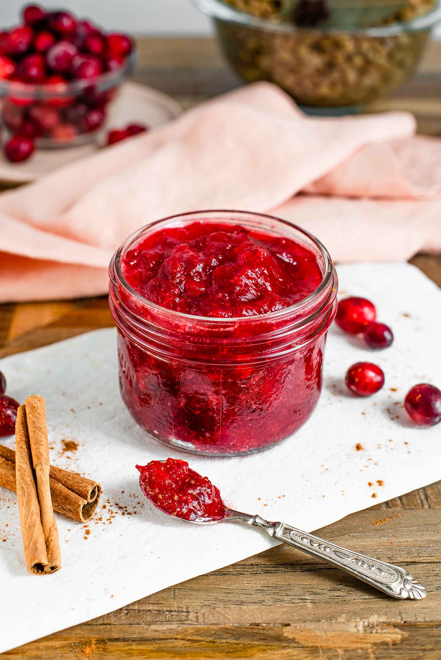 Side view of a jar of jammy cranberry sauce. A spoon with the thick sauce lays on a white tray in front of the jar. The tray is speckled with ground cinnamon, cinnamon sticks lay to one side, and fresh cranberries are scattered. 