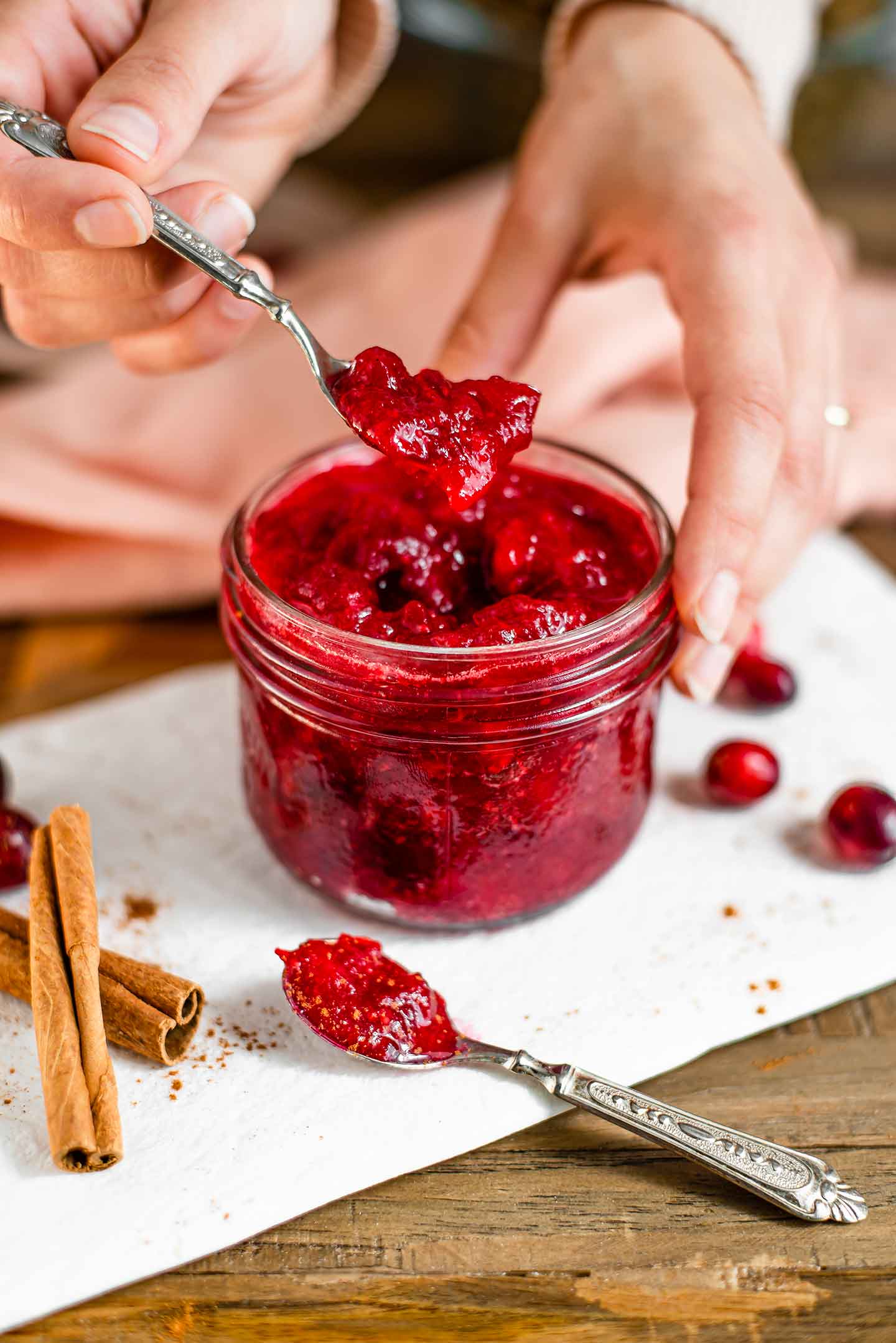 Side view of a hand lifting a spoon of cranberry sauce from a jar full of the red jam. Cinnamon sticks and fresh cranberries lay on a white tray surrounding the jar.