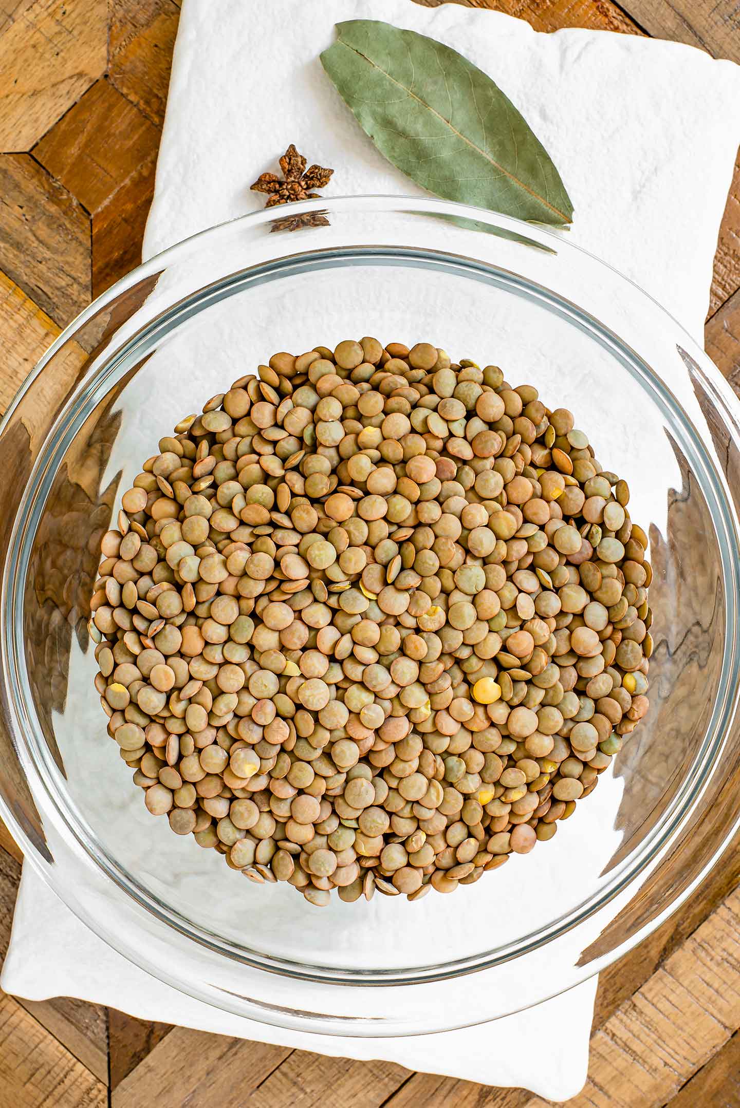Top down view of dried green lentils in a glass bowl atop a white tray. A bay leaf and star anise pod lay on the white tray.