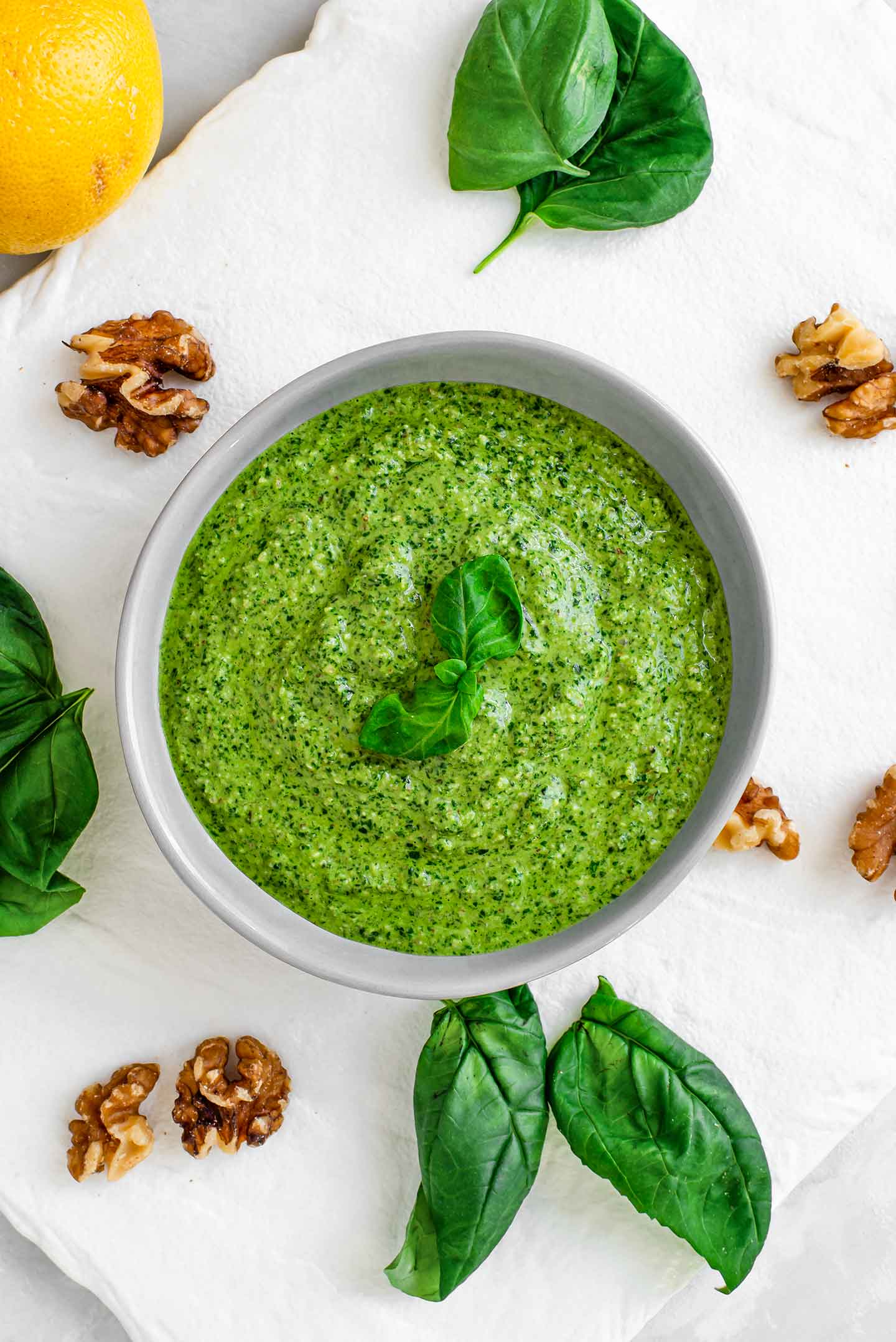 Top down view of homemade basil pesto in a bowl. Walnuts, basil leaves and a lemon are scattered around the pesto.