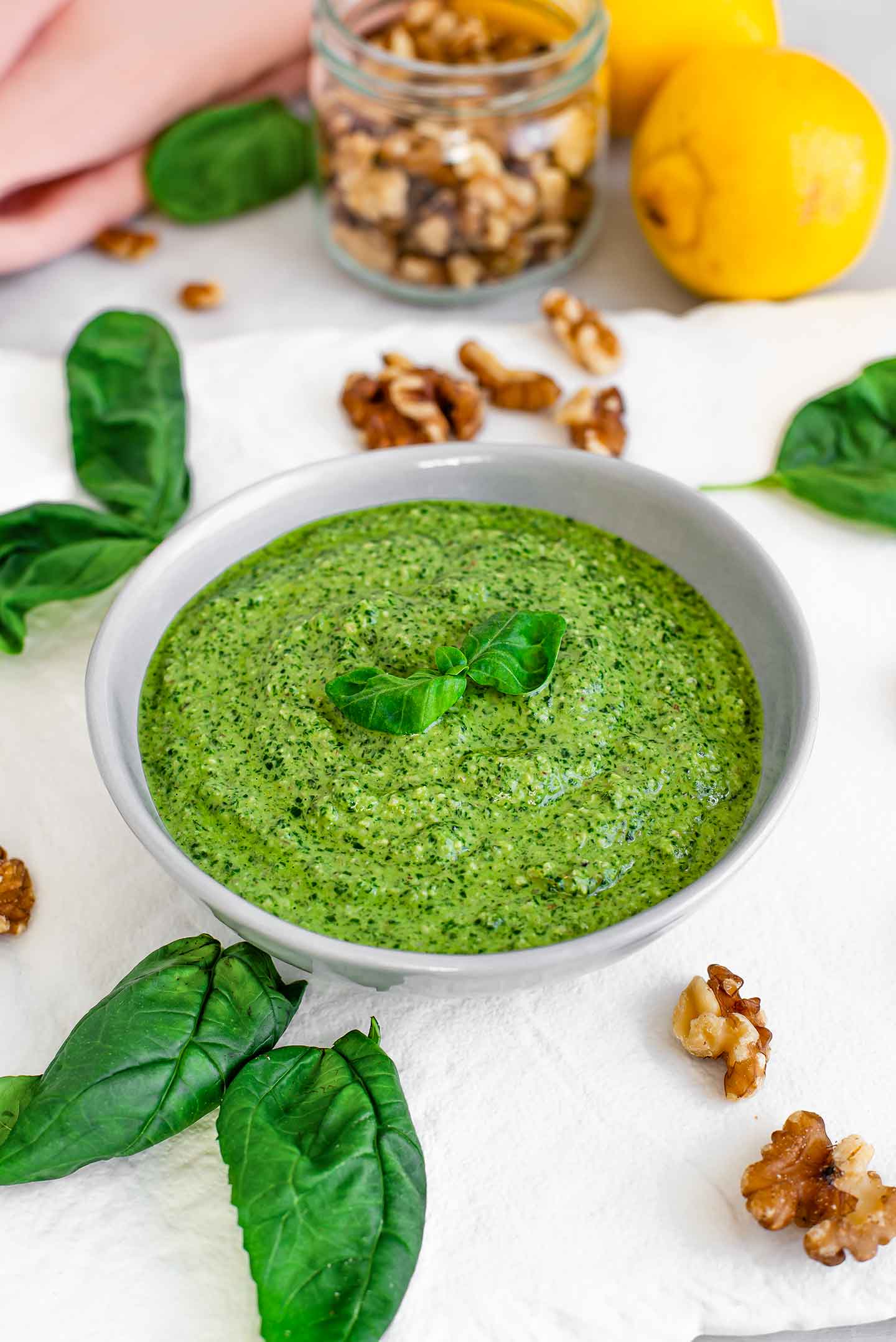Side view of basil pesto in a dish with small basil leaves garnishing the top. Walnuts, more basil, and a lemon surround the dish.