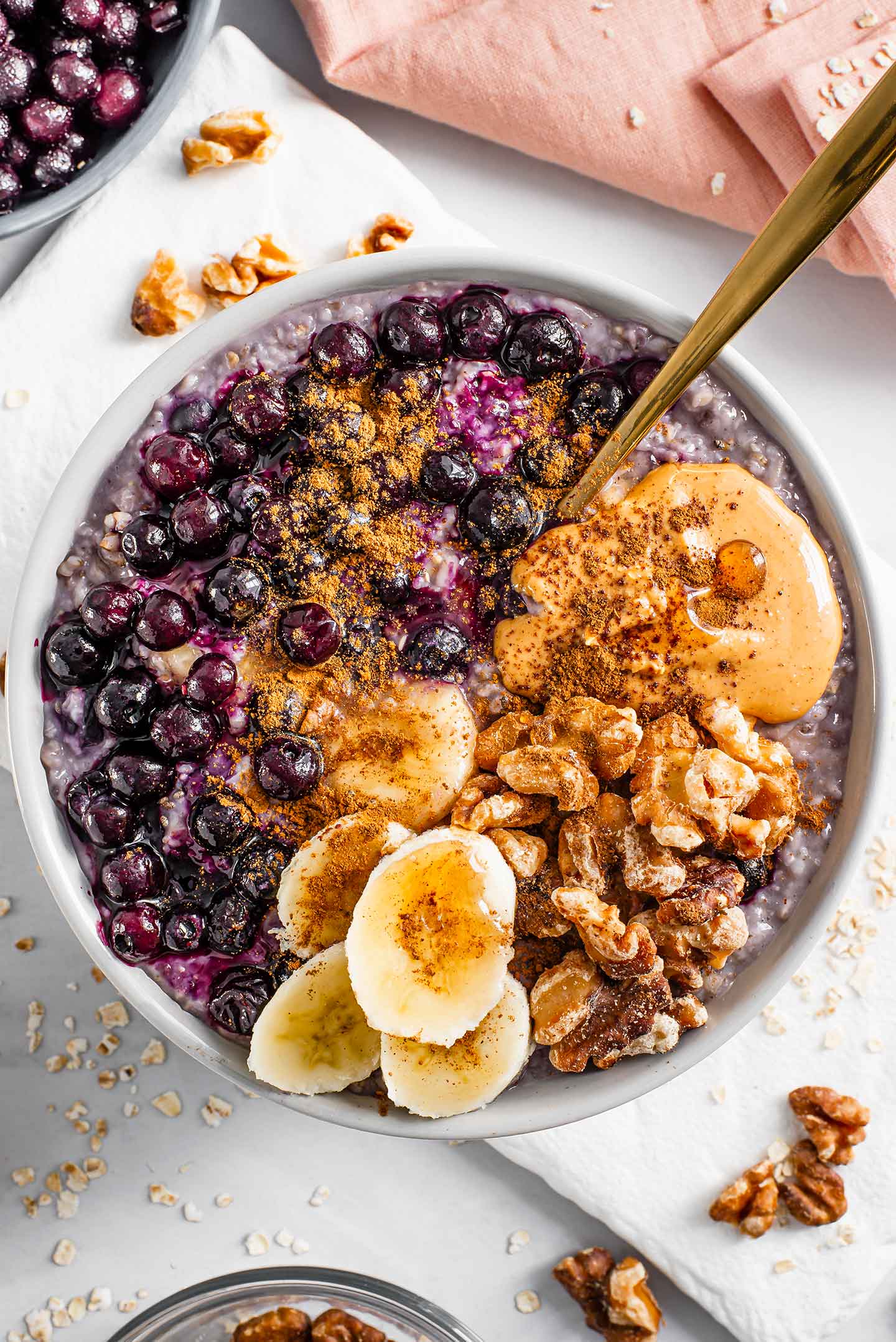 Top down view of blueberry banana oatmeal loaded with toppings. Saucy blueberries, banana slices, toasted walnuts, a dollop of peanut butter, and a sprinkle of cinnamon fill the top of the oats.
