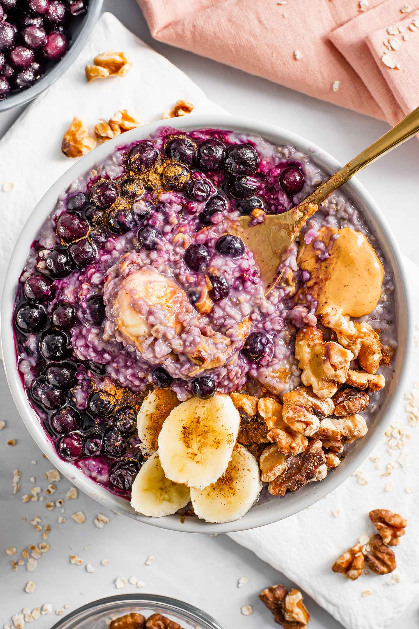 Top down view of a spoon scooping up purple oats from a loaded bowl of blueberry banana oatmeal. The top is decorated with extra blueberries, peanut butter, sliced banana, and toasted walnuts.