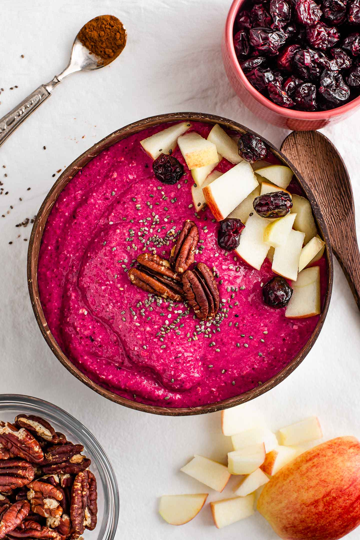 A bright pink smoothie bowl topped with diced apple, toasted pecans, chia seeds, and dried cranberries.