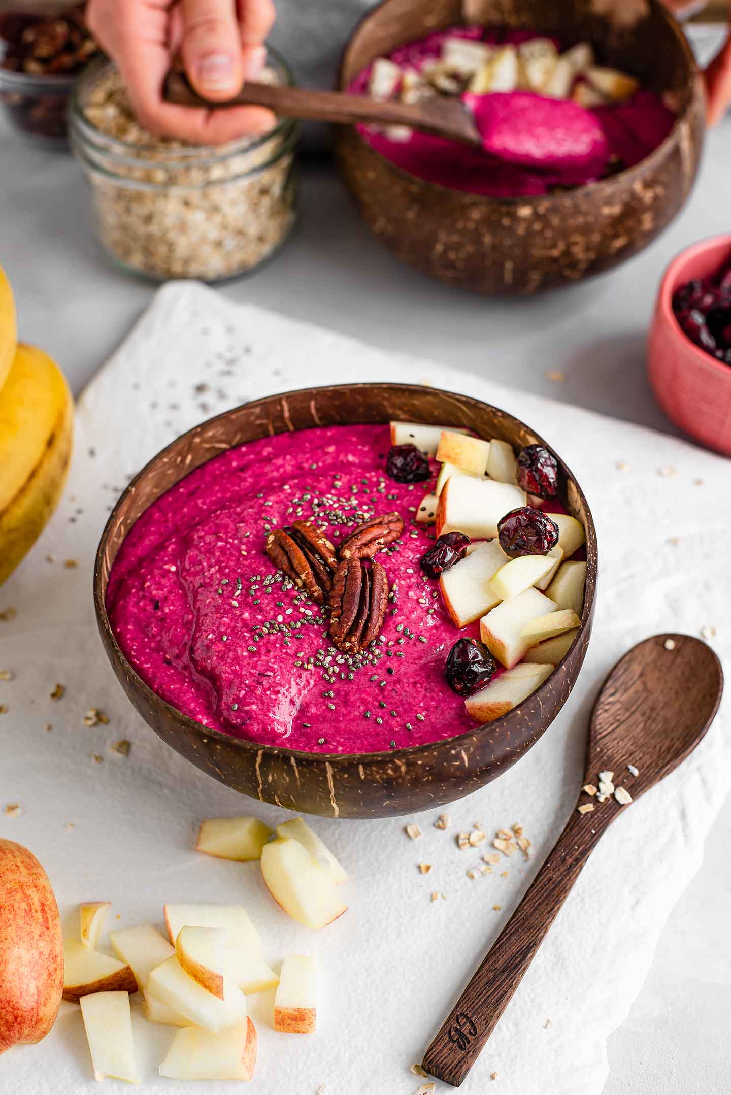 Top down view of a beet apple smoothie bowl with a wooden spoon laying next to it on a white tray. Ingredients are scattered around.