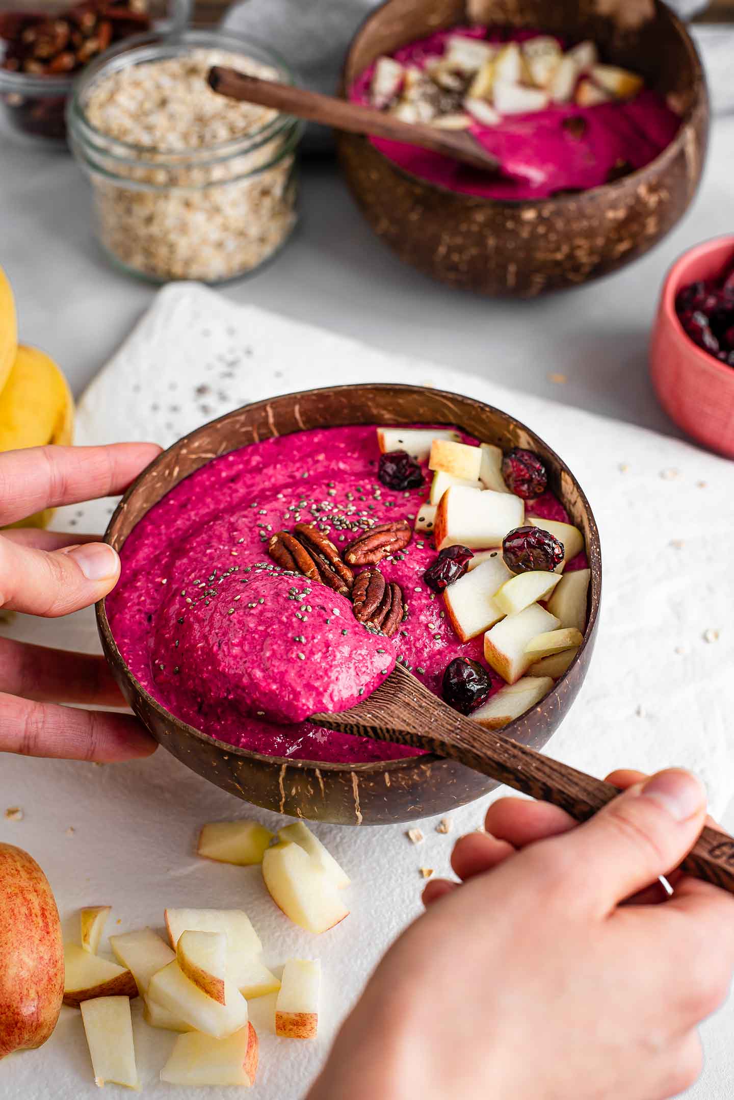 Side view of a hand using a wooden spoon to scoop the hot pink apple beet smoothie from the bowl. Another smoothie, oats, diced apple, cranberries, and toasted pecans are in the background.
