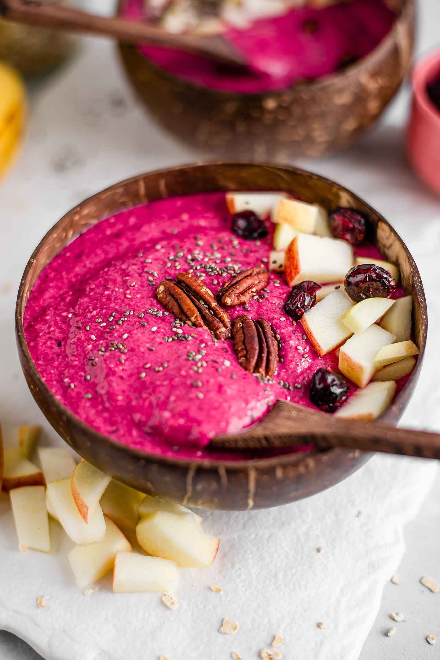 Side view of a wooden spoon dipped into a thick hot pink cinnamon spiced apple beet smoothie bowl topped with diced apple, dried cranberries, toasted pecans, and chia seeds.