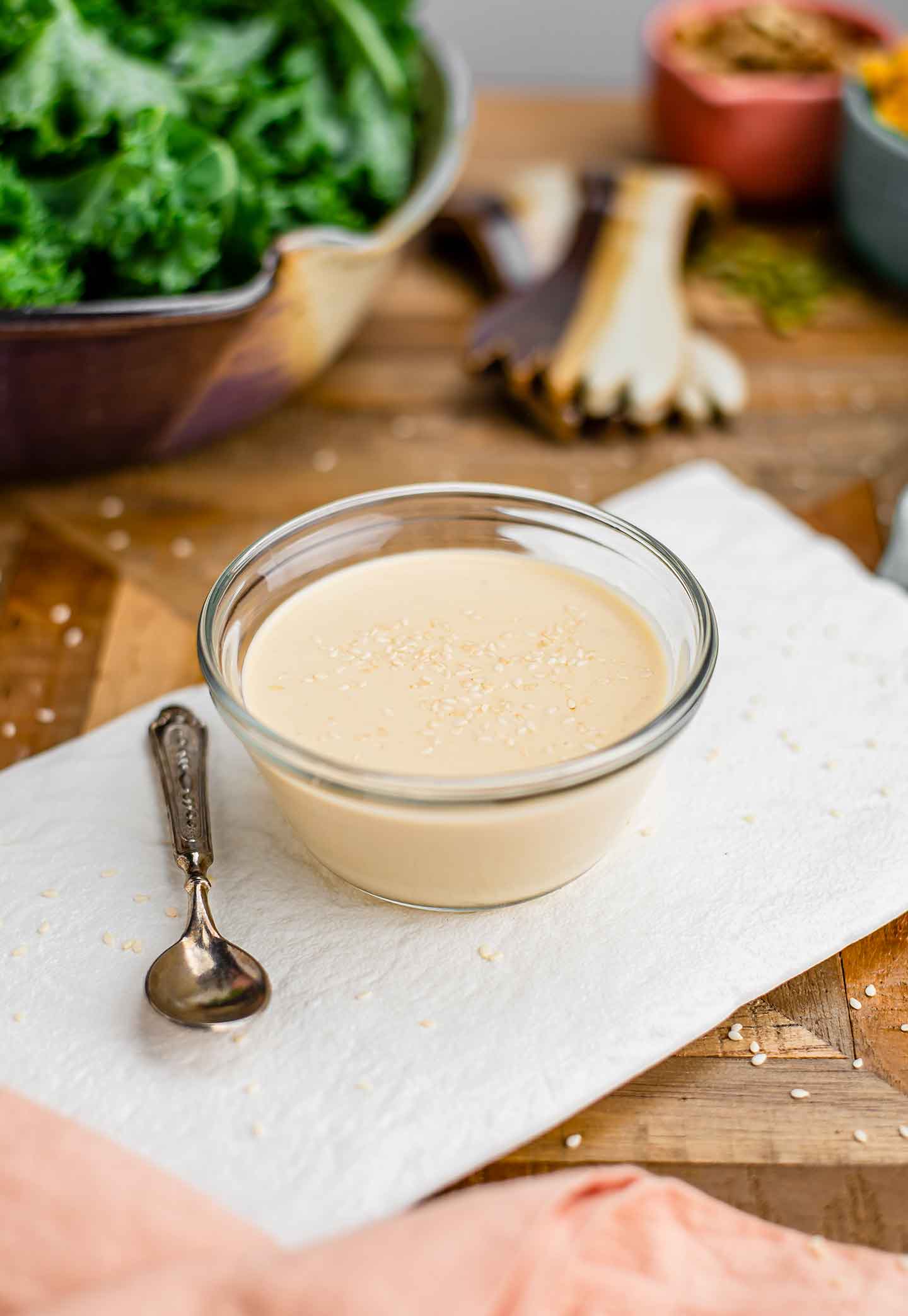 Side view of a thick lemon tahini dressing garnished with sesame seeds filling a small bowl on a white tray. Kale leaves and ingredients for making a salad fill the background.