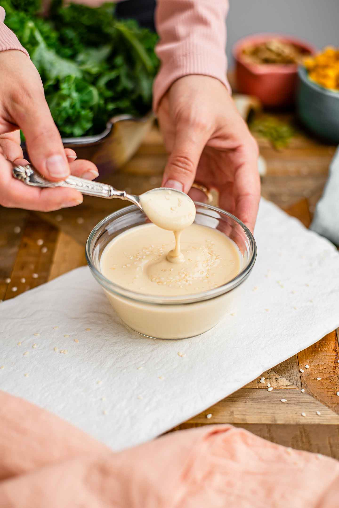Side view of a hand lifting a spoon of creamy lemon tahini dressing from a small bowl. The dressing is thick but pourable and garnished with sesame seeds.