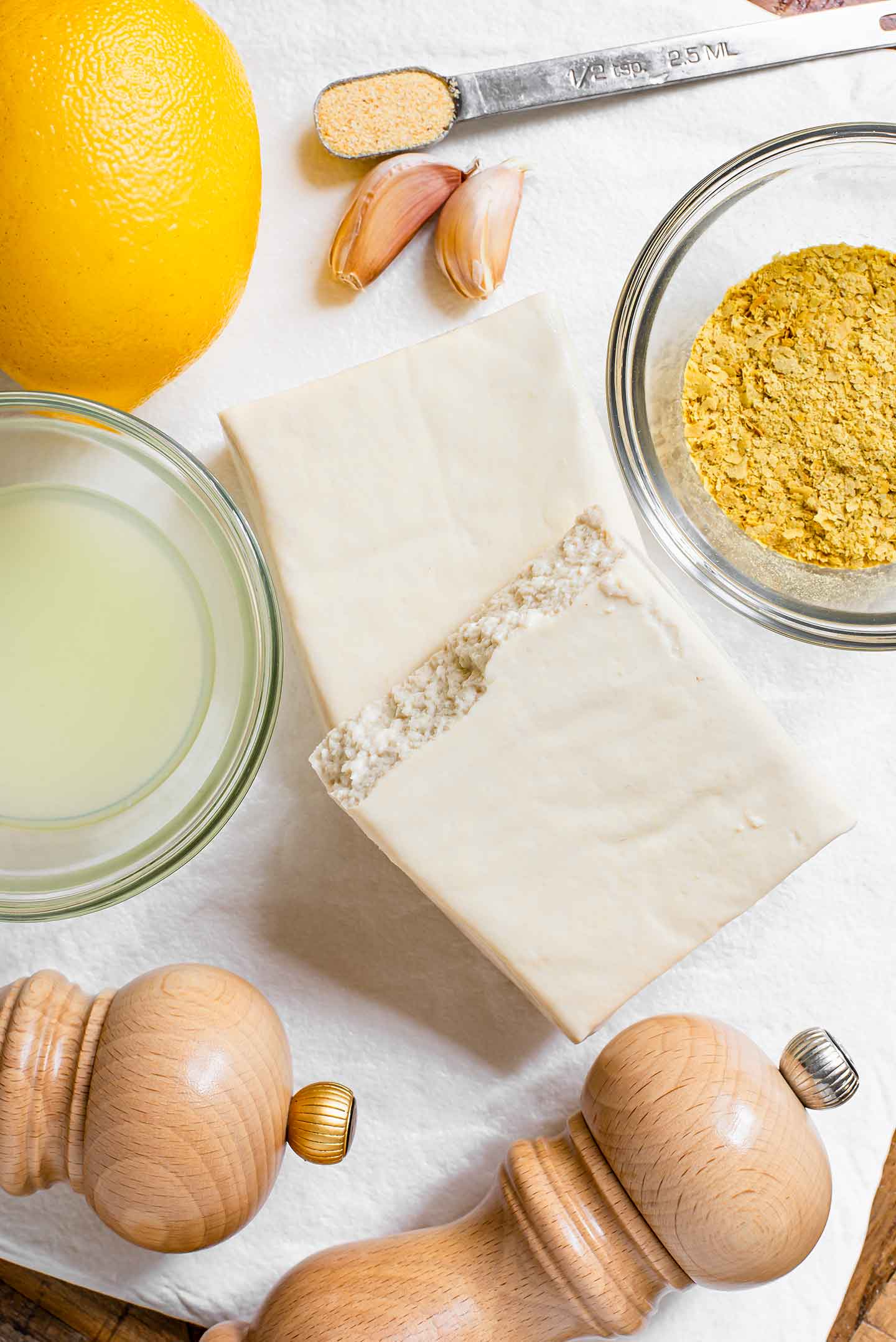Top down view of ingredients on a white tray. a block of extra firm tofu is torn in half and surrounded by a lemon, garlic cloves, nutritional yeast, a dish of lemon juice and salt and pepper mills.