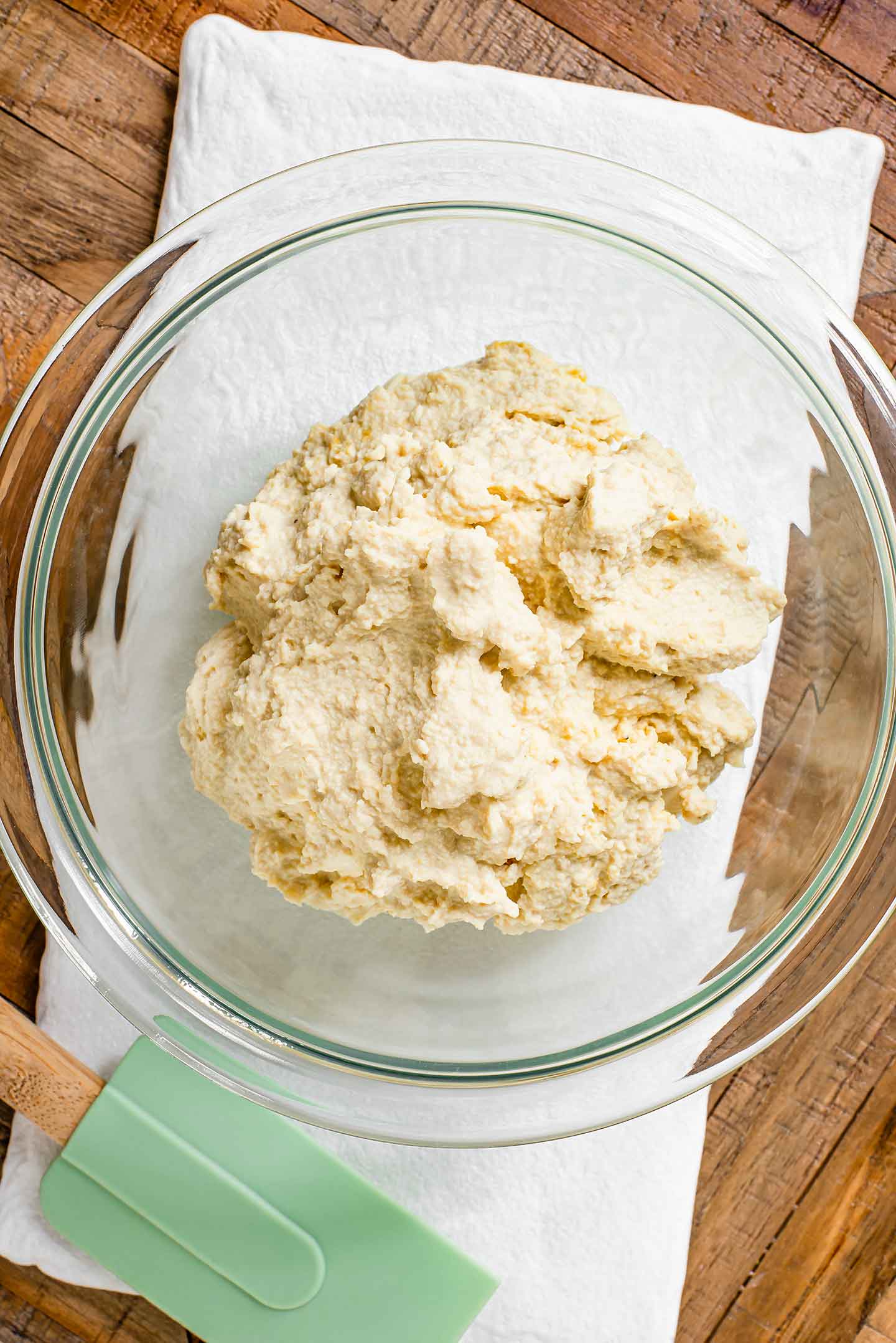 Top down view of textured tofu ricotta in a glass bowl resting on a white tray. A light green rubber spatula lays next to the bowl.