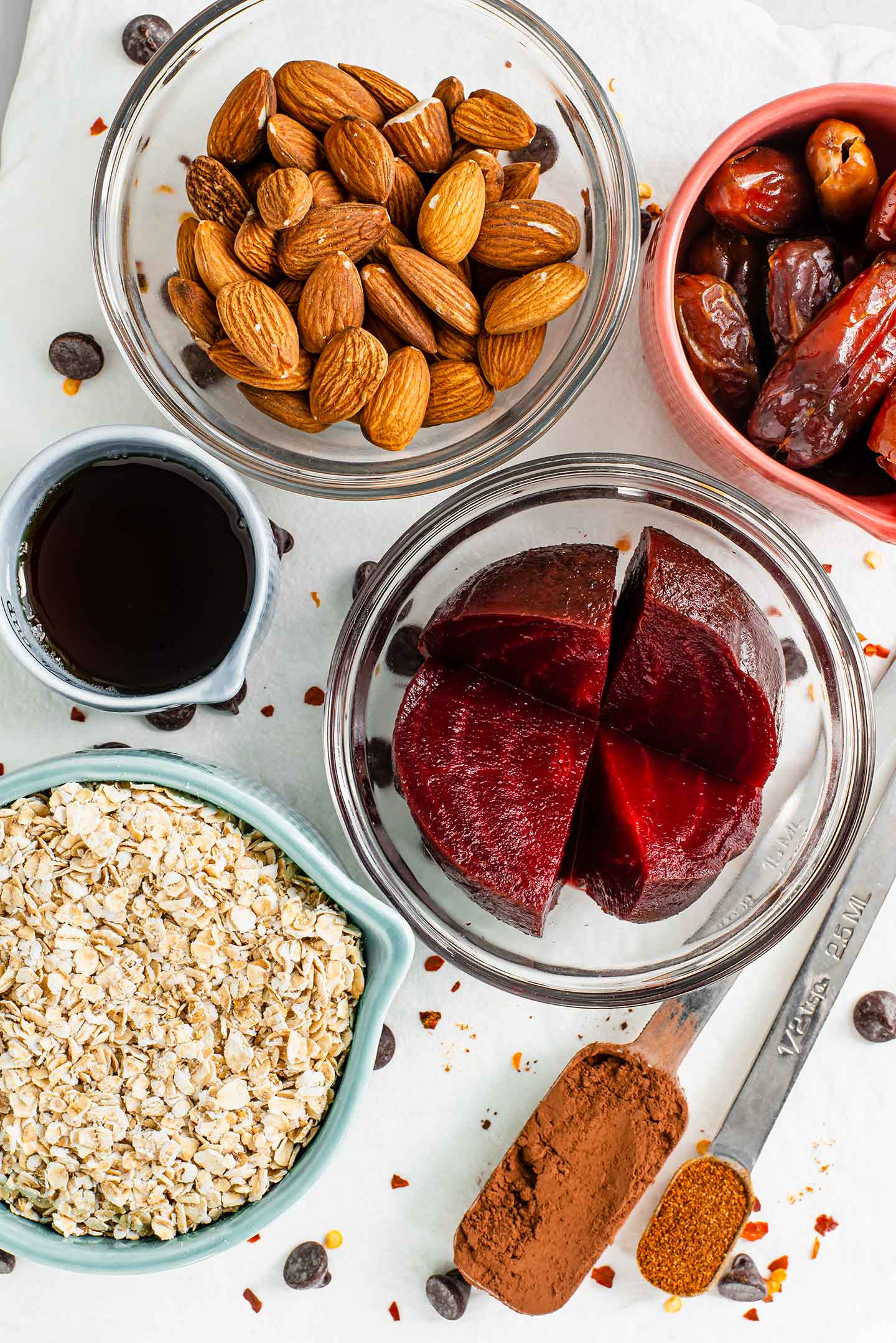Top down view of ingredients in separate dishes. Red beets, almonds, dates, oats, maple syrup, cocoa powder, chili powder, and chocolate chips. 