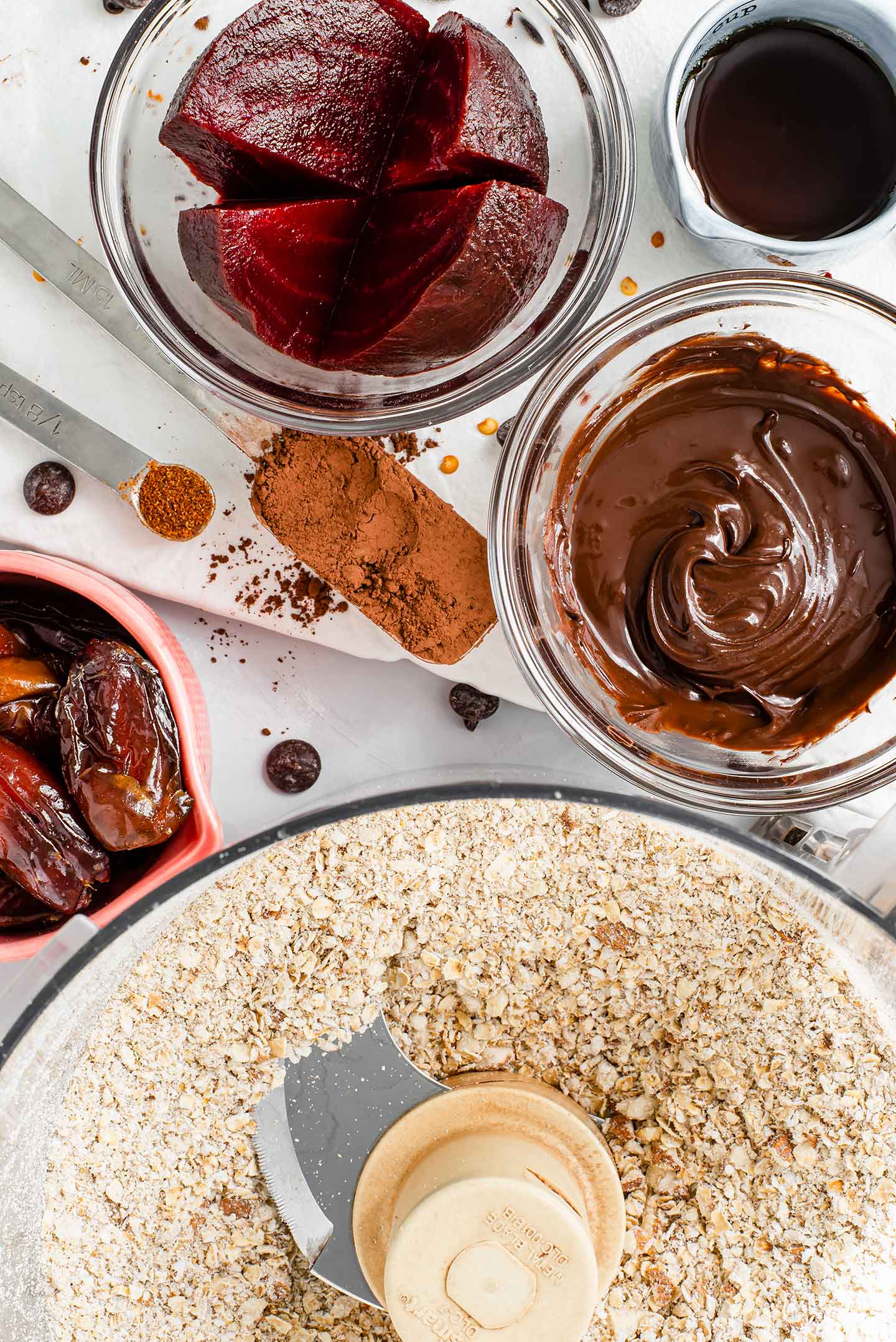 Top down view of oats and almonds ground in a food processor with dishes of melted chocolate chips, sliced beets, whole dates, and maple syrup waiting to be added to the mixture. 