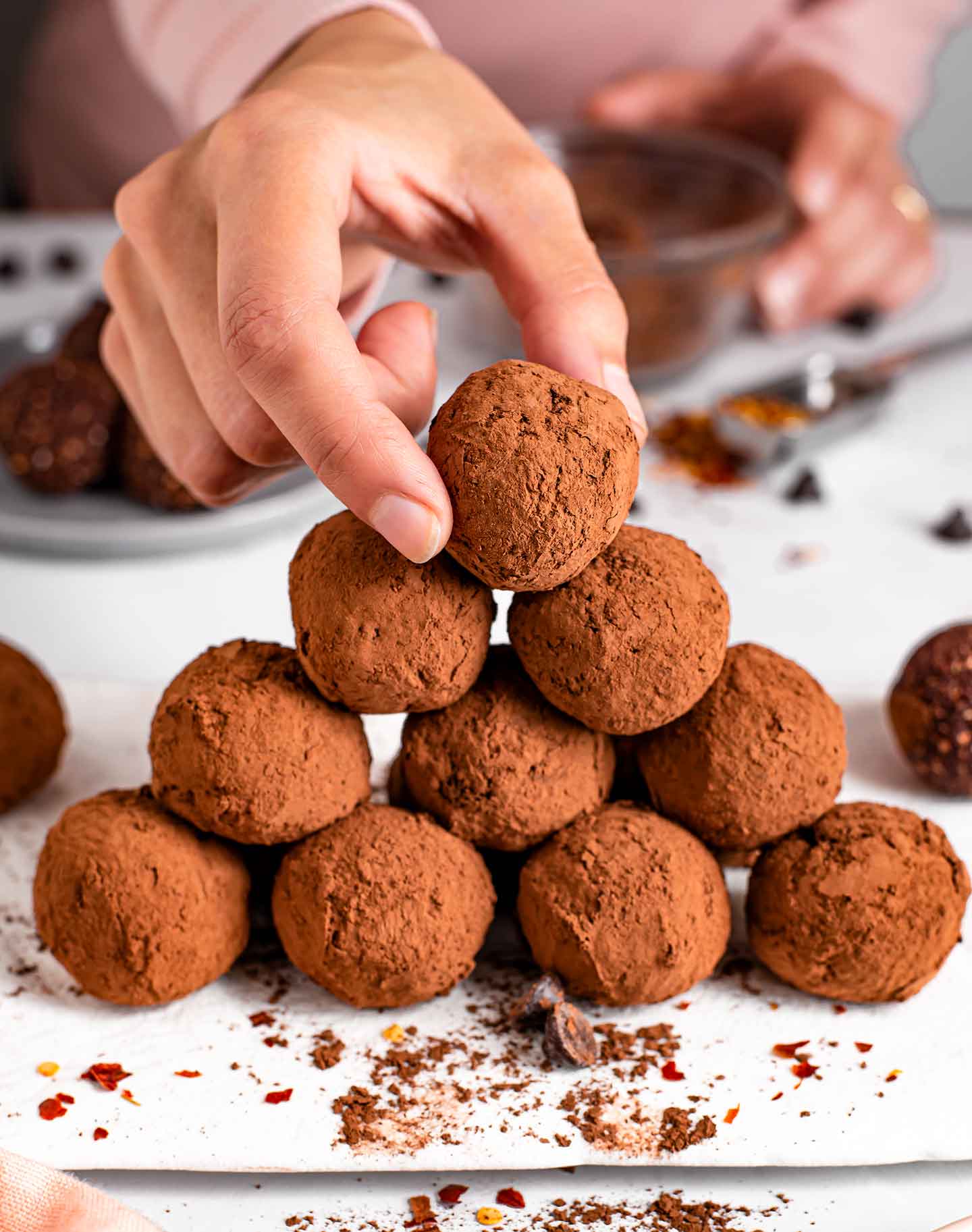 Side view of a diamond shaped stack of truffle chili energy balls. Fingers reach fro the top ball. Cocoa powder and chocolate chips are scattered around.
