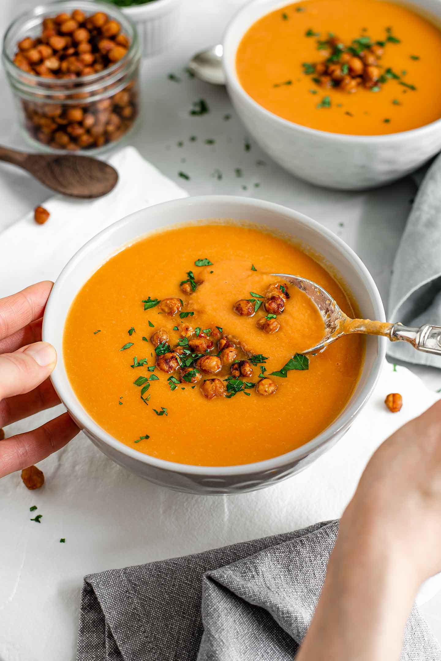 Side view of a hand dipping a spoon into the bright orange, creamy soup. The soup is thick and topped with crispy roasted chickpeas.