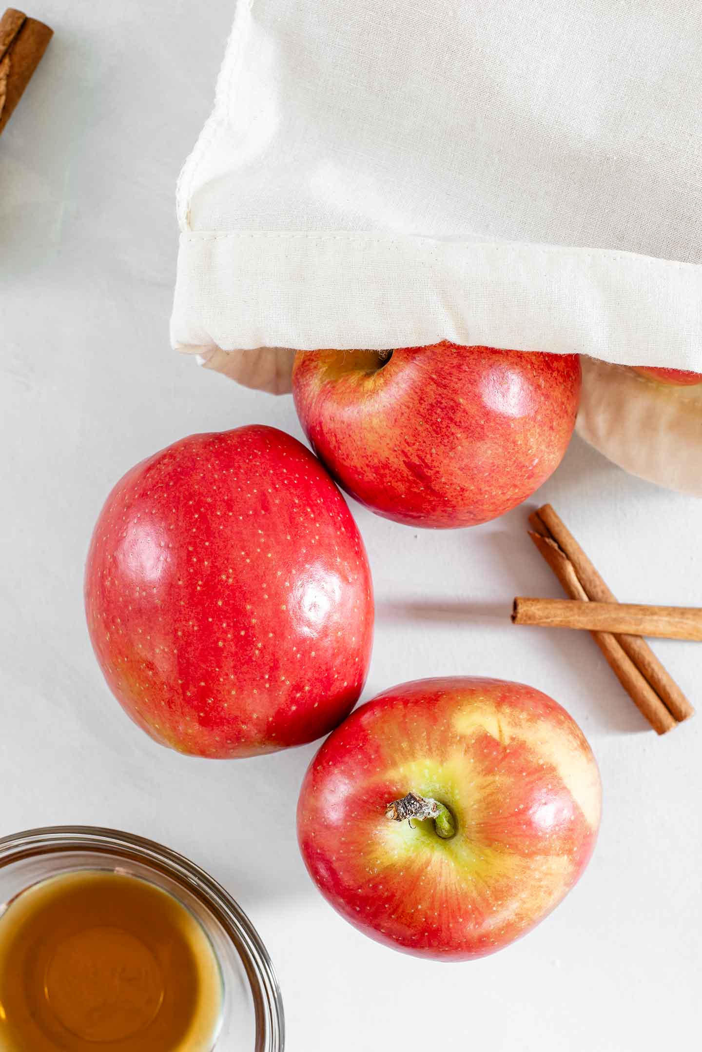 Top down view of apples spilling out of a canvas produce bag with cinnamon sticks and a small glass bowl of maple syrup to the side