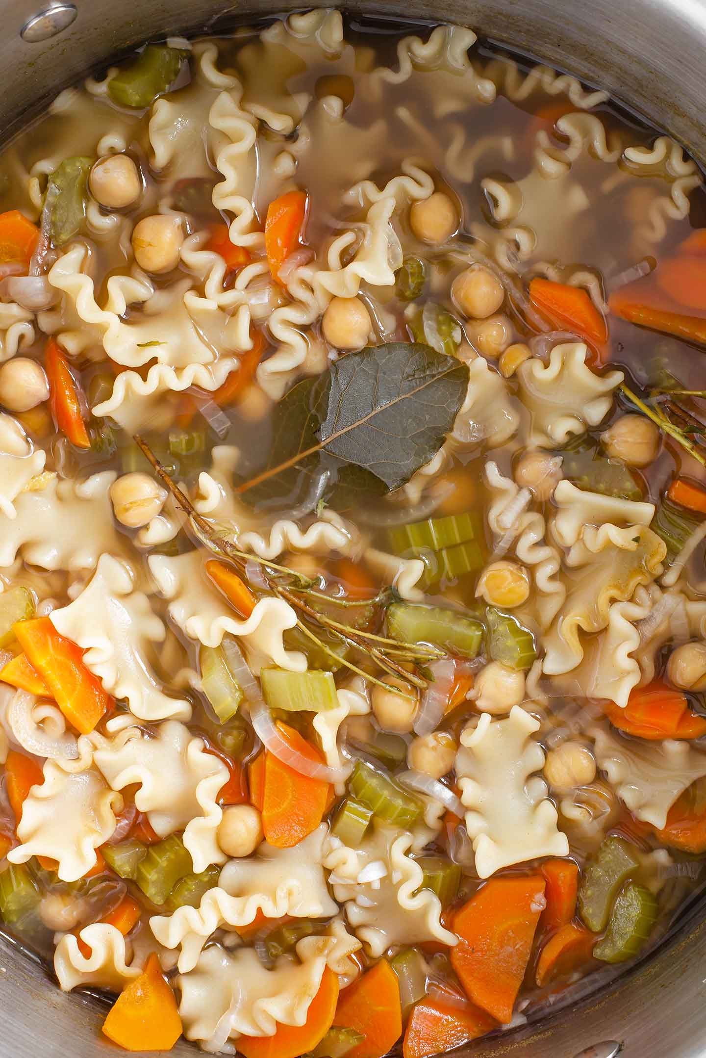 Top down view of easy noodle soup cooked in a pot. The bay leaf and thyme sprigs float in the broth, on top of thick noodles, diced carrots, chopped celery, and chickpeas.