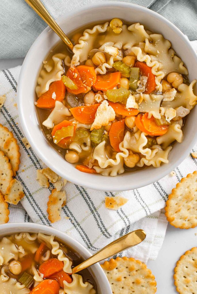 Top down view of easy noodle soup in a bowl with a spoon and crumbled crackers on top. The soup is loaded with carrots, celery, chickpeas, and thick mini lasagna noodles. Another bowl of soup is visible in the corner and crackers are scattered around.