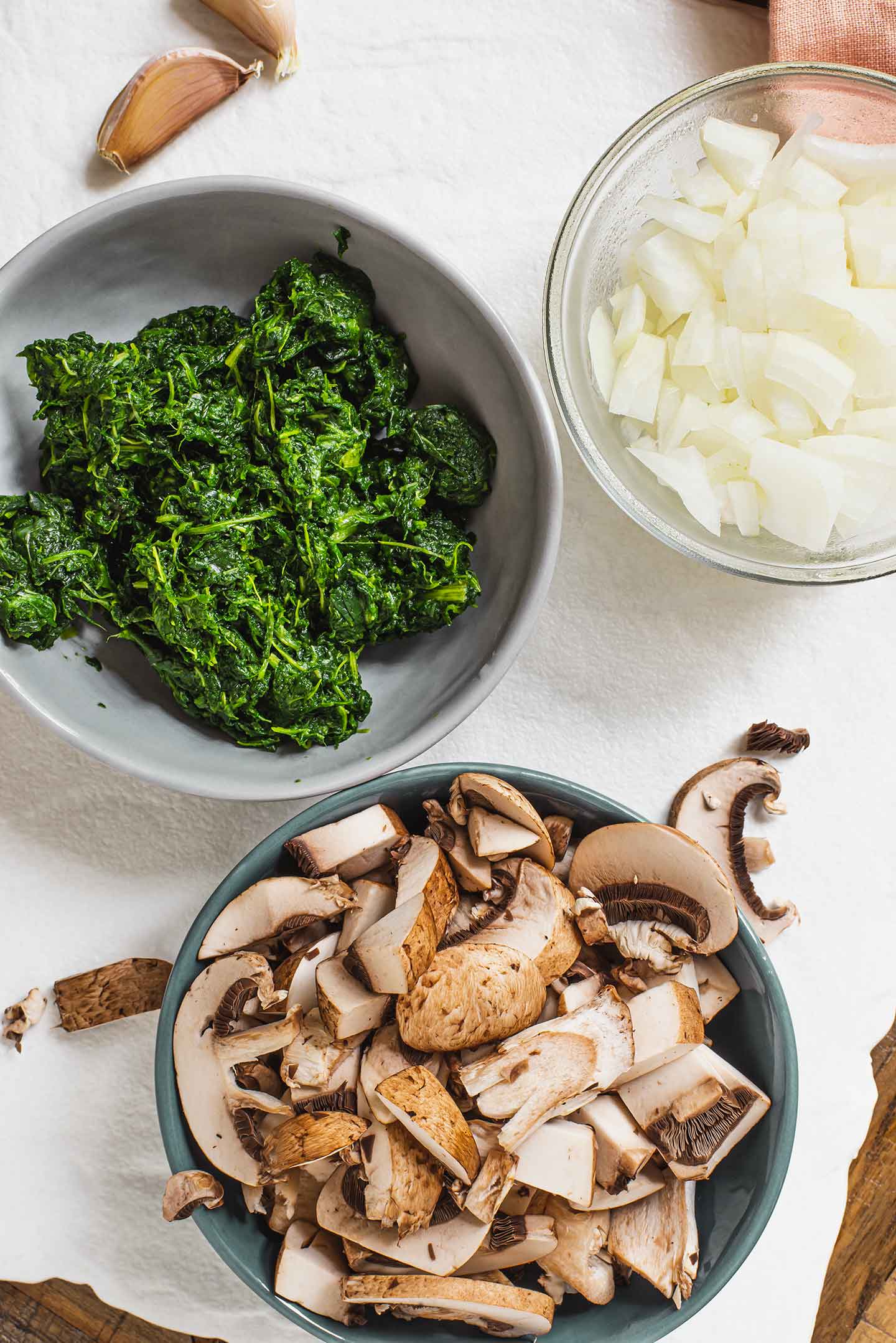 Top down view of ingredients atop a white tray. Sliced mushrooms, cooked spinach, diced onion, and garlic cloves.