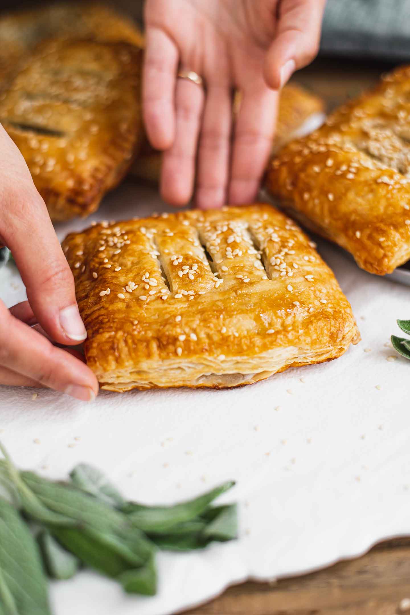 Side view of two hands laying a vegan puff pastry pocket on a white tray. The side of the pastry is visible and is nicely laminated, browned, and flaky.