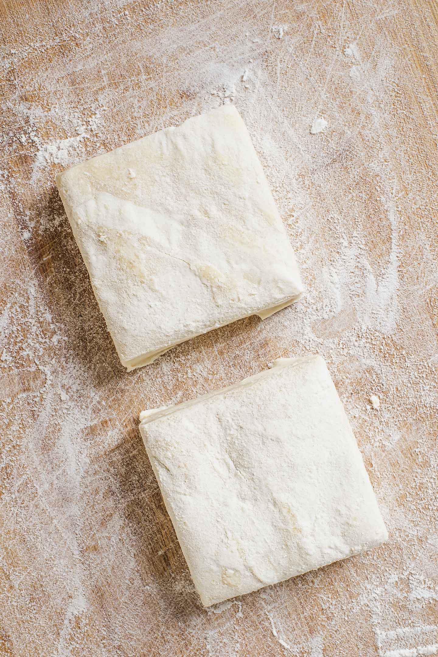 Top down view of two squares of puff pastry on a lightly floured wooden cutting board.