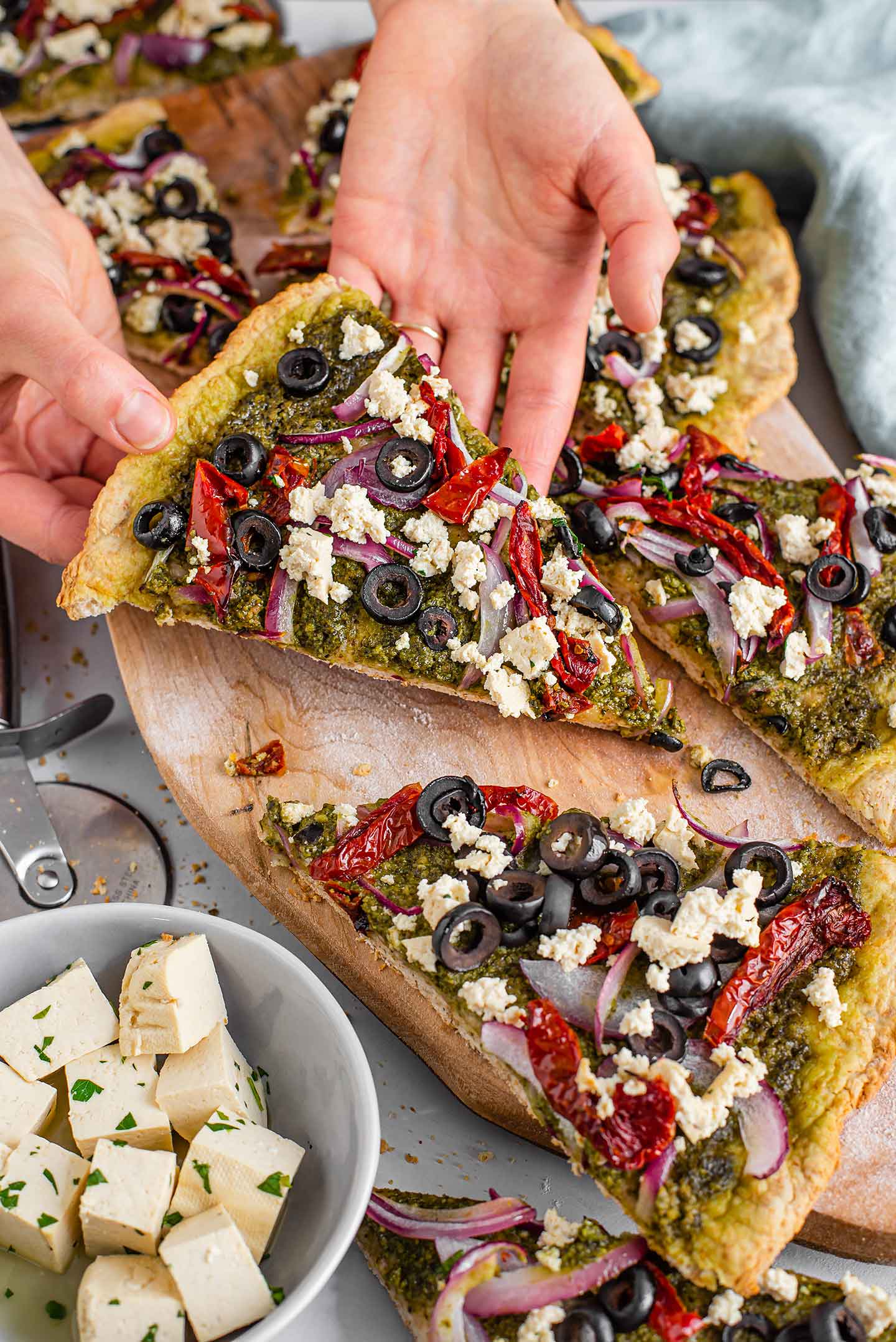 Top down view of a hand lifting a slice of thin crust pesto pizza with feta crumble from a wooden tray. Extra feta fills a small dish and other slices are scattered around.