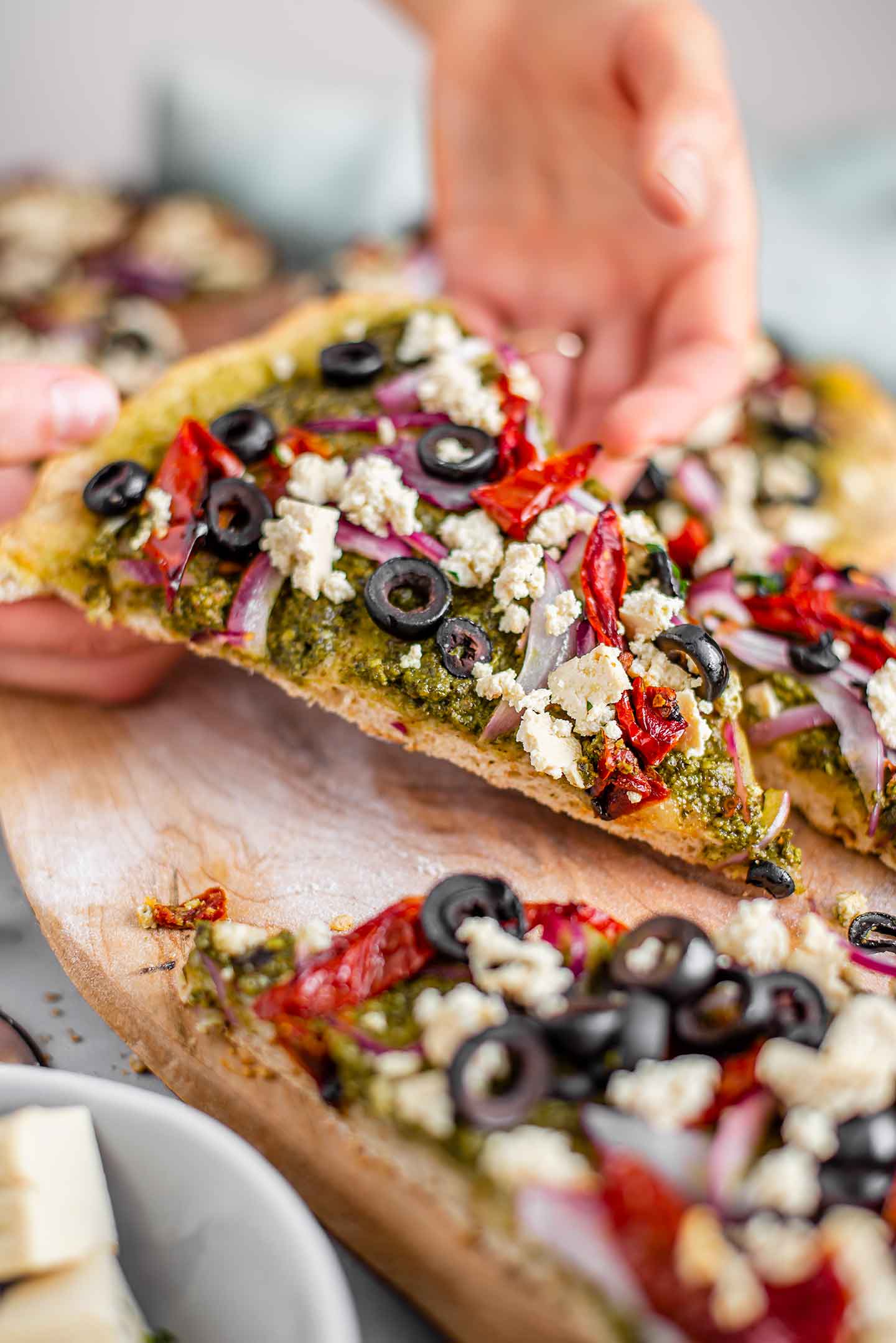 Side view of hand lifting a slice of pesto pizza with feta from a wooden tray.