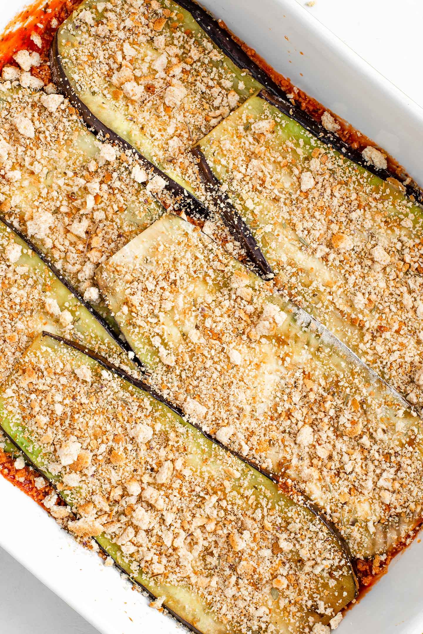 Top down view of sliced eggplant sprinkled with breadcrumbs in a casserole dish. The eggplant sits atop a thin layer of tomato sauce.