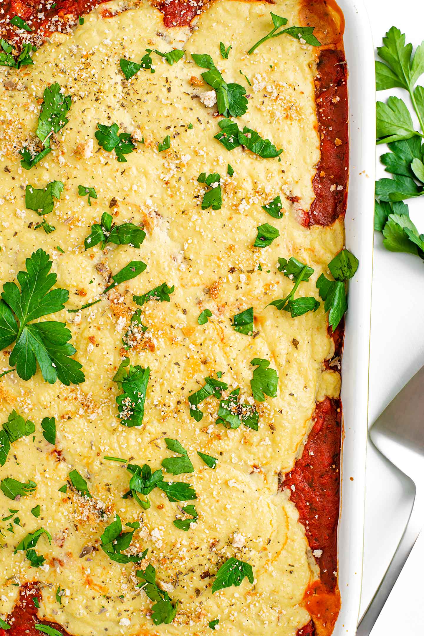 Top down view of vegan baked eggplant "parmesan" in a casserole dish. Parsley sits atop golden bechamel. Tomato sauce peaks through underneath and a spatula lays next to the casserole dish.