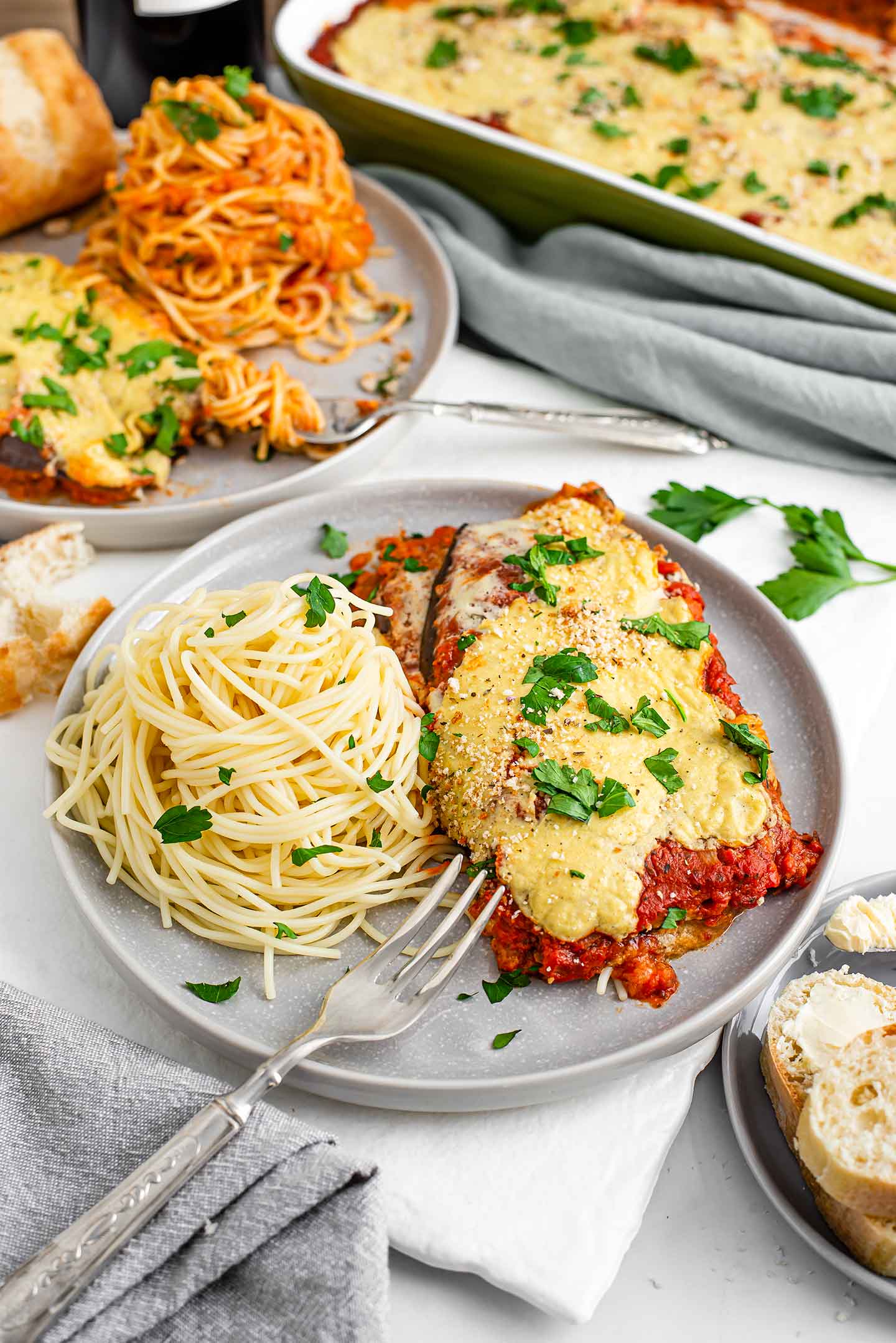 A saucy and cheesy slice of vegan baked eggplant "parm" with a mound of spaghetti beside it and a fork resting on the plate. 