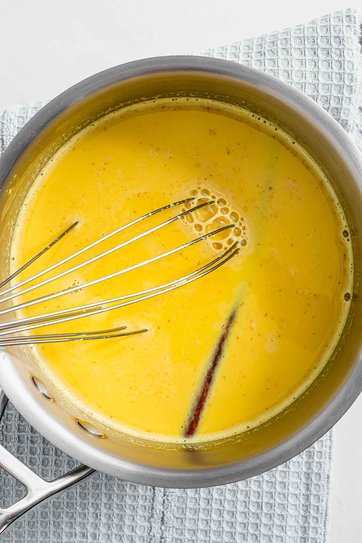 Top down view of golden milk simmering in a pot. A cinnamon stick is visible through the vibrant yellow drink and a whisk sits in the centre.