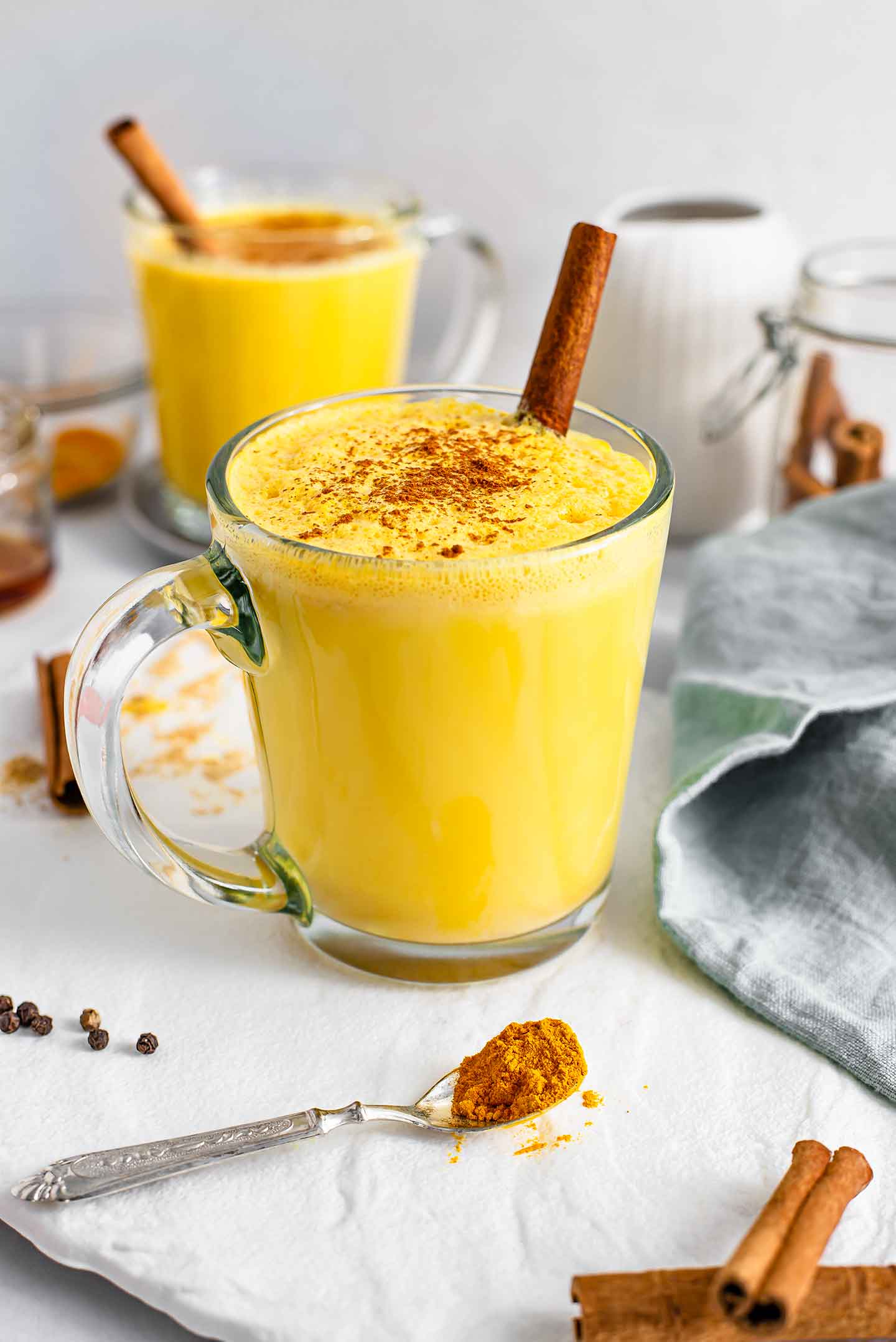 Golden turmeric milk fills a clear glass mug. A cinnamon stick protrudes from the foamy top and ground cinnamon is sprinkled on top. Another mug of milk, turmeric powder, and cinnamon sticks are in the background.