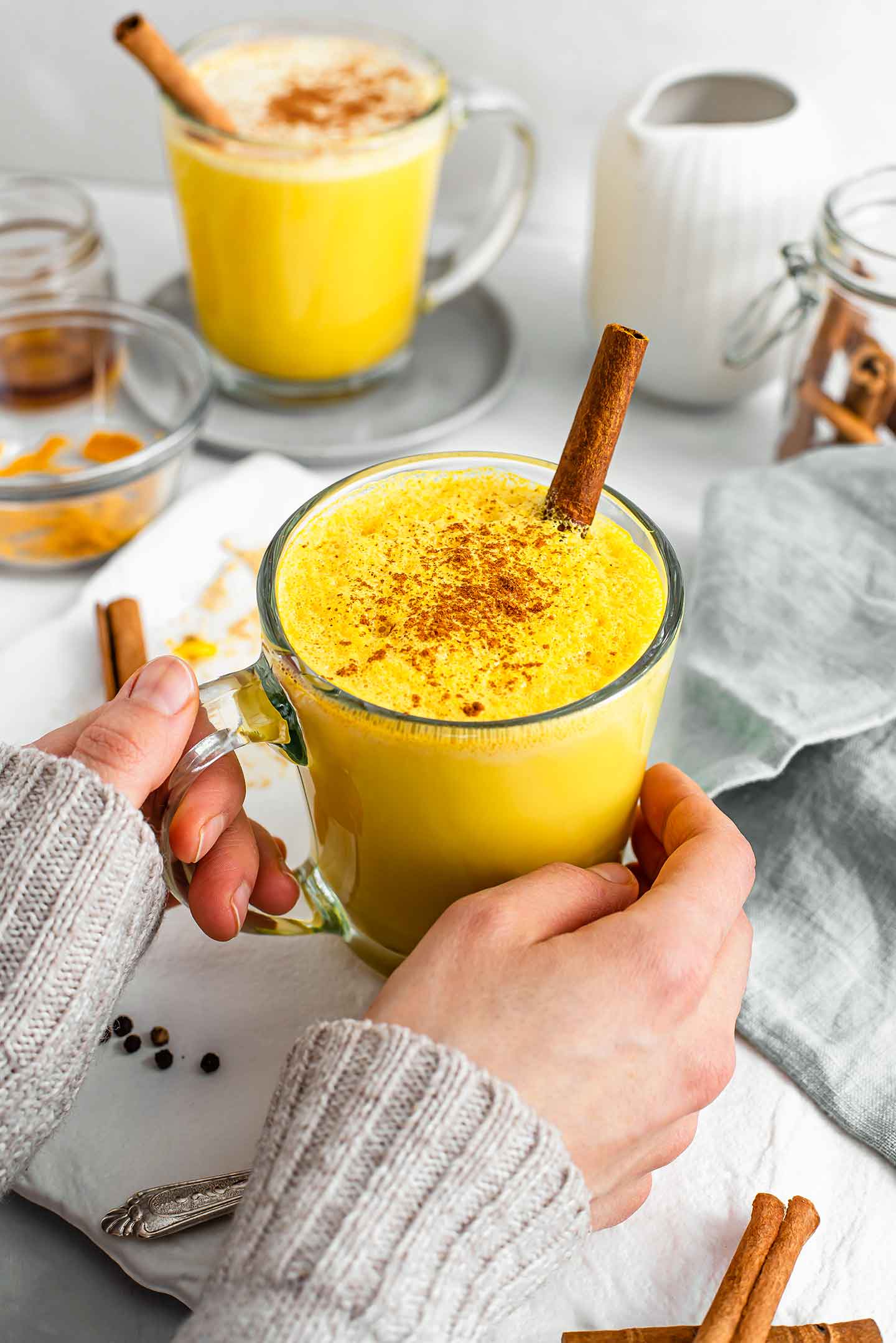 Two hands with a cozy sweater visible, rest on the sides of the clear mug. The latte is vibrant yellow with a cinnamon stick protruding. Another latte and ingredients are in the background.