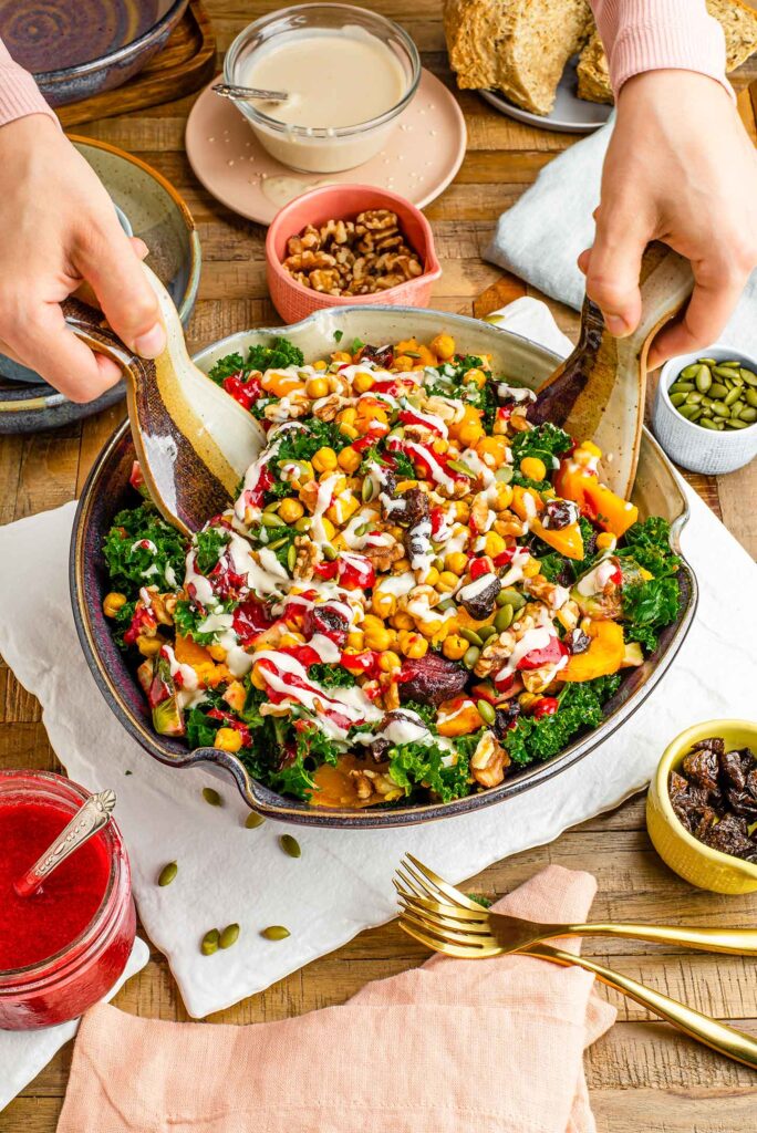 Top down view of a large colourful salad in a ceramic bowl with two hands tossing the salad with ceramic tongs. A creamy dressing and cranberry sauce are drizzled on the top.