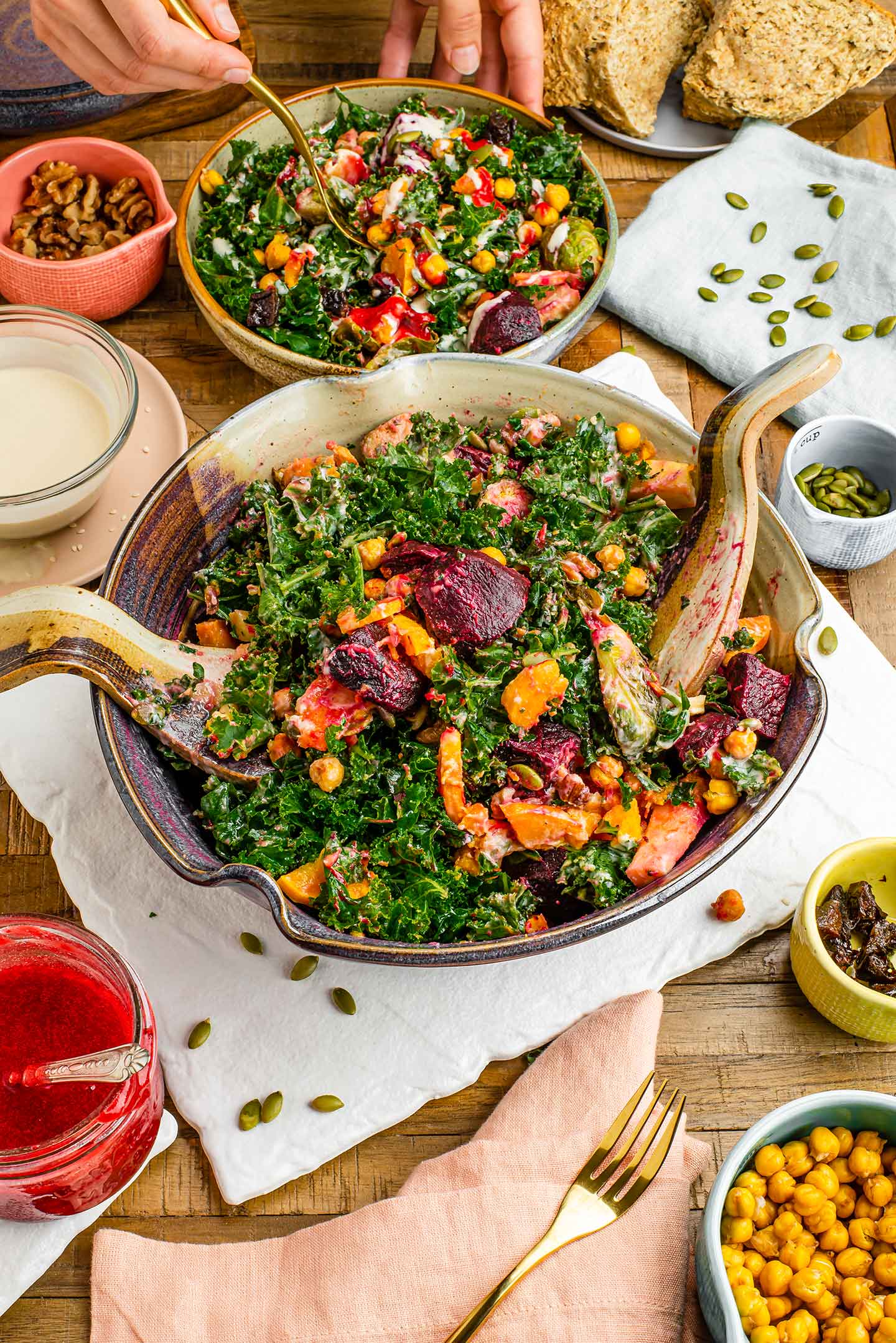 Top down view of a kale salad with roasted vegetables tossed and portioned into a smaller bowl. Ceramic tongs sit in the large salad bowl and pumpkin seeds are sprinkled around.